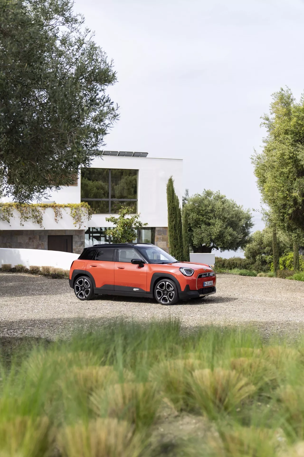 2024 Mini Aceman (J05) four-door compact SUV on gravel, showcasing front and side view with contemporary house and greenery backdrop, featuring contrast-colored roof, modern LED headlights, and potential high-end customization