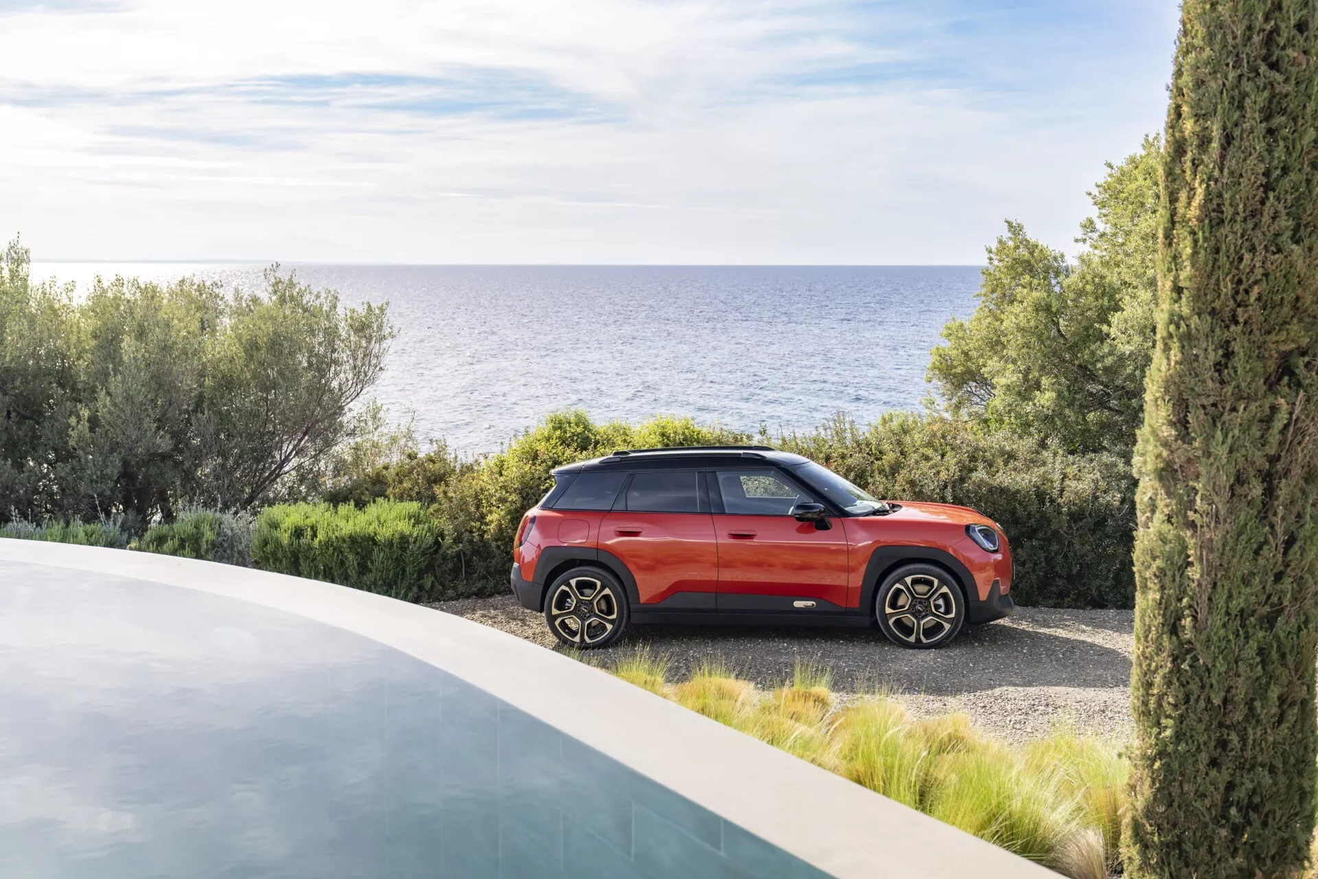 2024 Mini Aceman (J05) parked near the coast, showcasing its modern crossover SUV design with a two-tone metallic paint finish, distinctive alloy wheels, and advanced lighting, set against a backdrop of a luxurious property and a serene sea view.