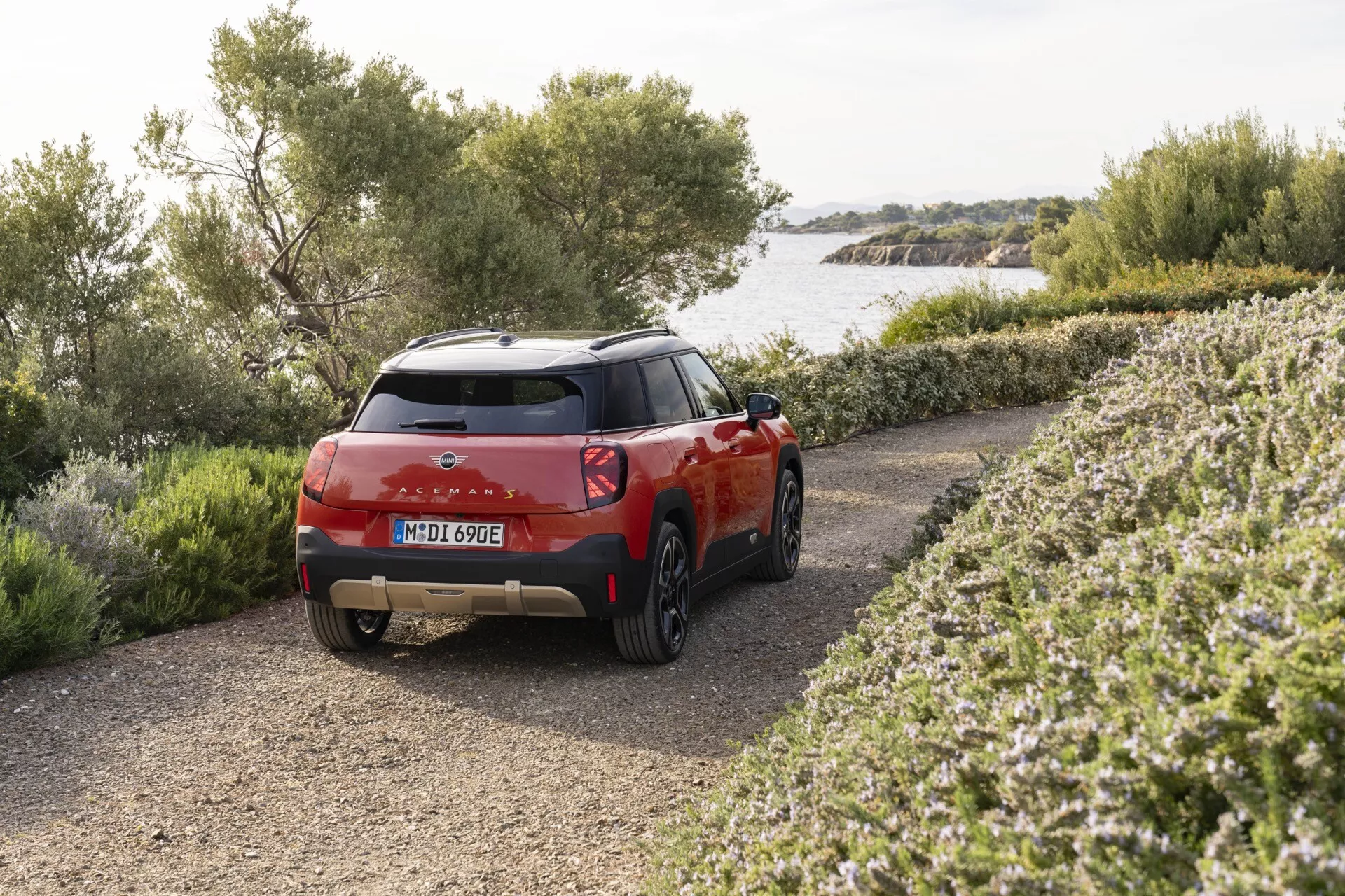 2024 Mini Aceman compact SUV in red parked outdoors showing three-quarter rear view highlighting LED taillights, dual-tone color scheme, and eco-friendly electric or hybrid model features against a rural or coastal backdrop with shrubbery and water