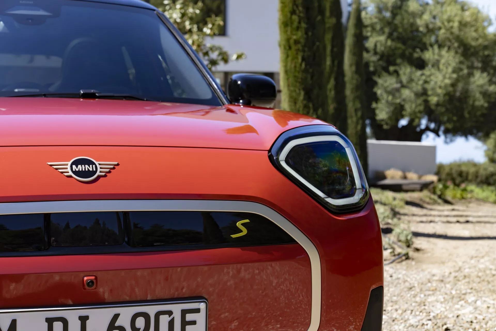 Close-up of a red 2024 Mini Aceman (J05) showcasing the sleek LED headlight, sporty grille with 'S' badge, and electric 'E' emblem on the front left side, reflecting its sporty, electric hybrid pedigree and luxurious design in bright outdoor light.