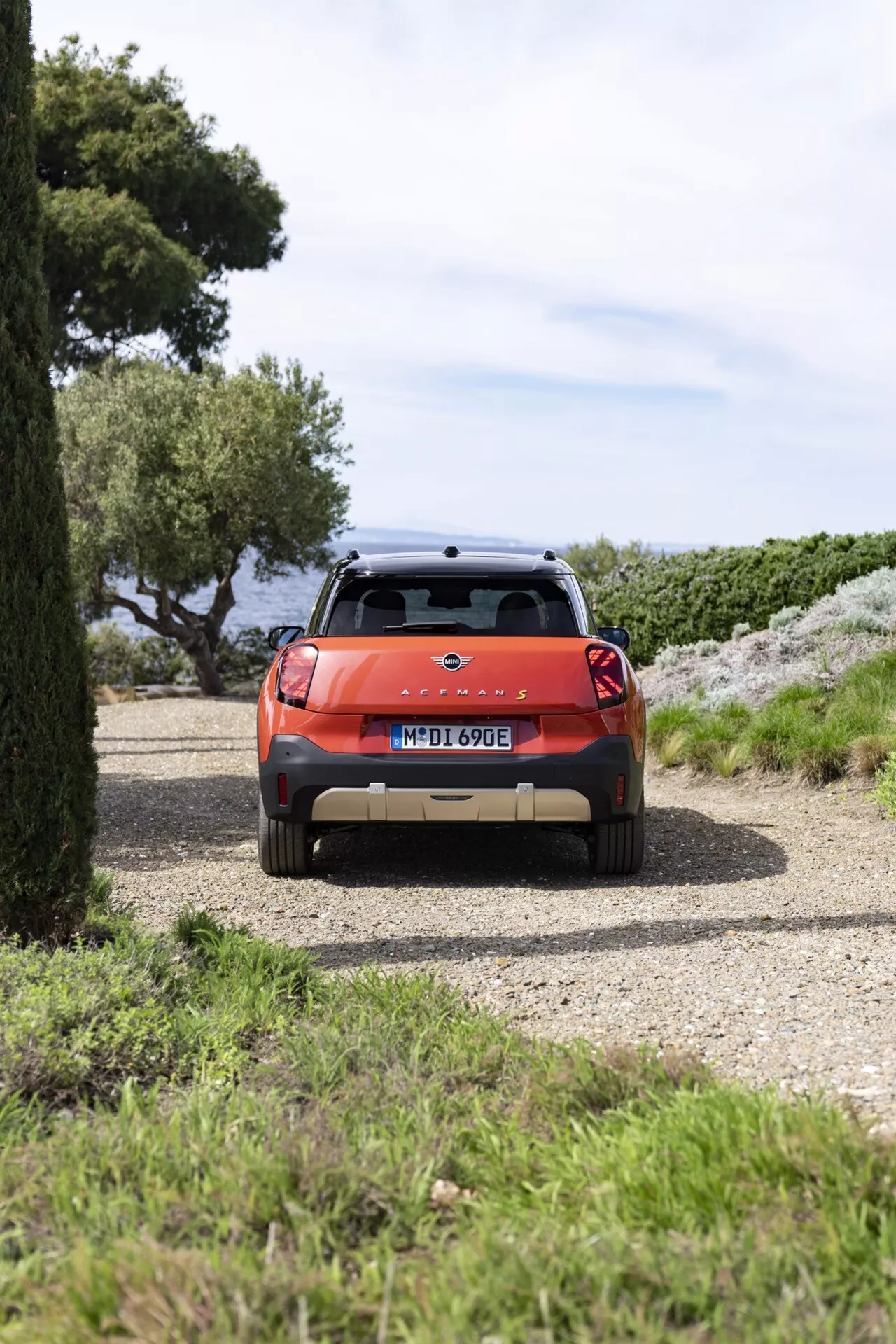 2024 Mini Aceman (J05) SUV in bright metallic orange, featuring taillights, rear windshield with wiper, license plate, roof spoiler with brake light, roof rails for extra cargo, parked on a gravel driveway with trees and possible ocean backdrop, showcasing its potential for adventure and outdoor activities