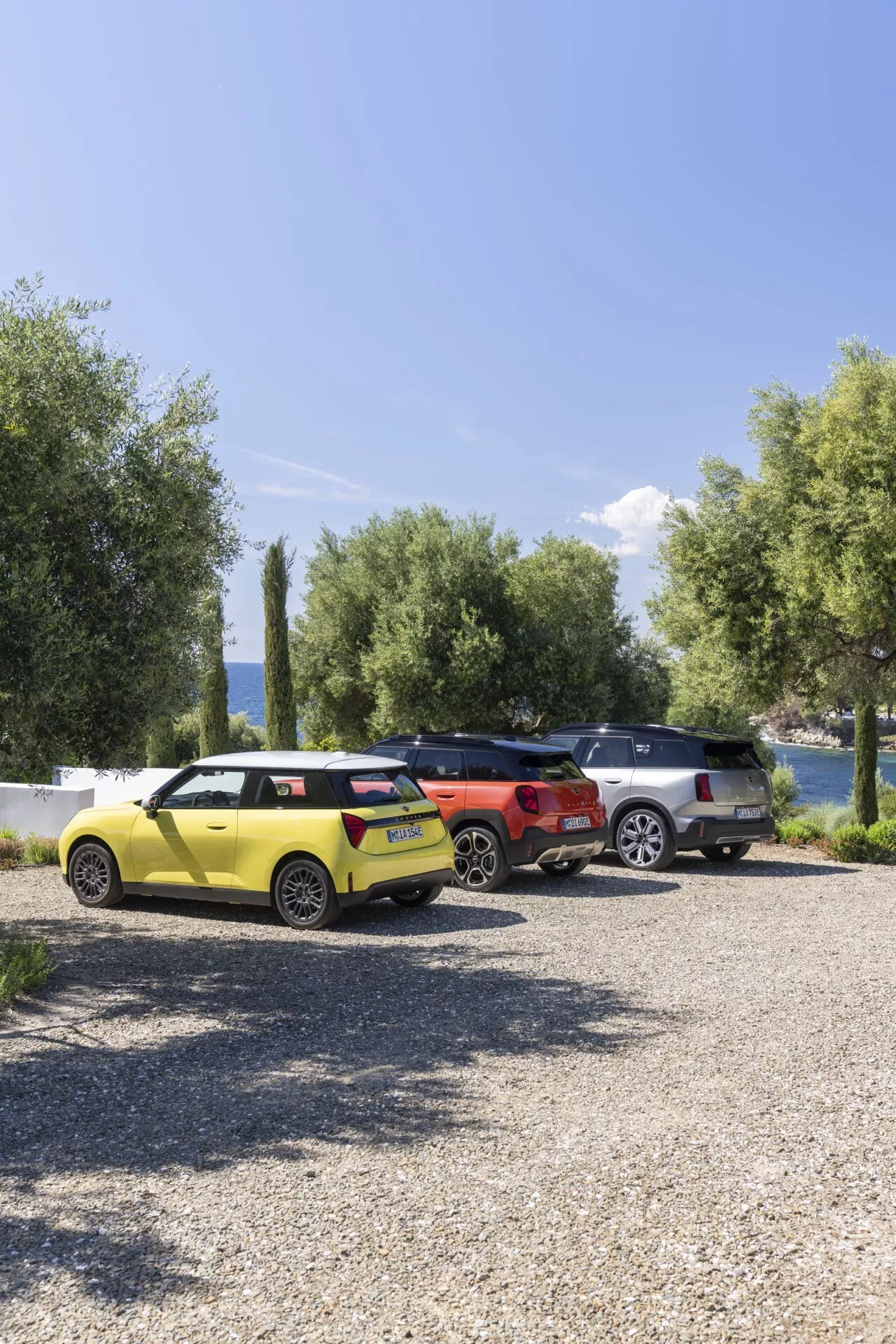 2024 Mini Aceman (J05) in bright yellow, orange, and grey, parked on a gravel surface with a natural backdrop consisting of trees, water, and the sky, showcasing modern design with sharp lines, LED taillights, and a compact to SUV size range
