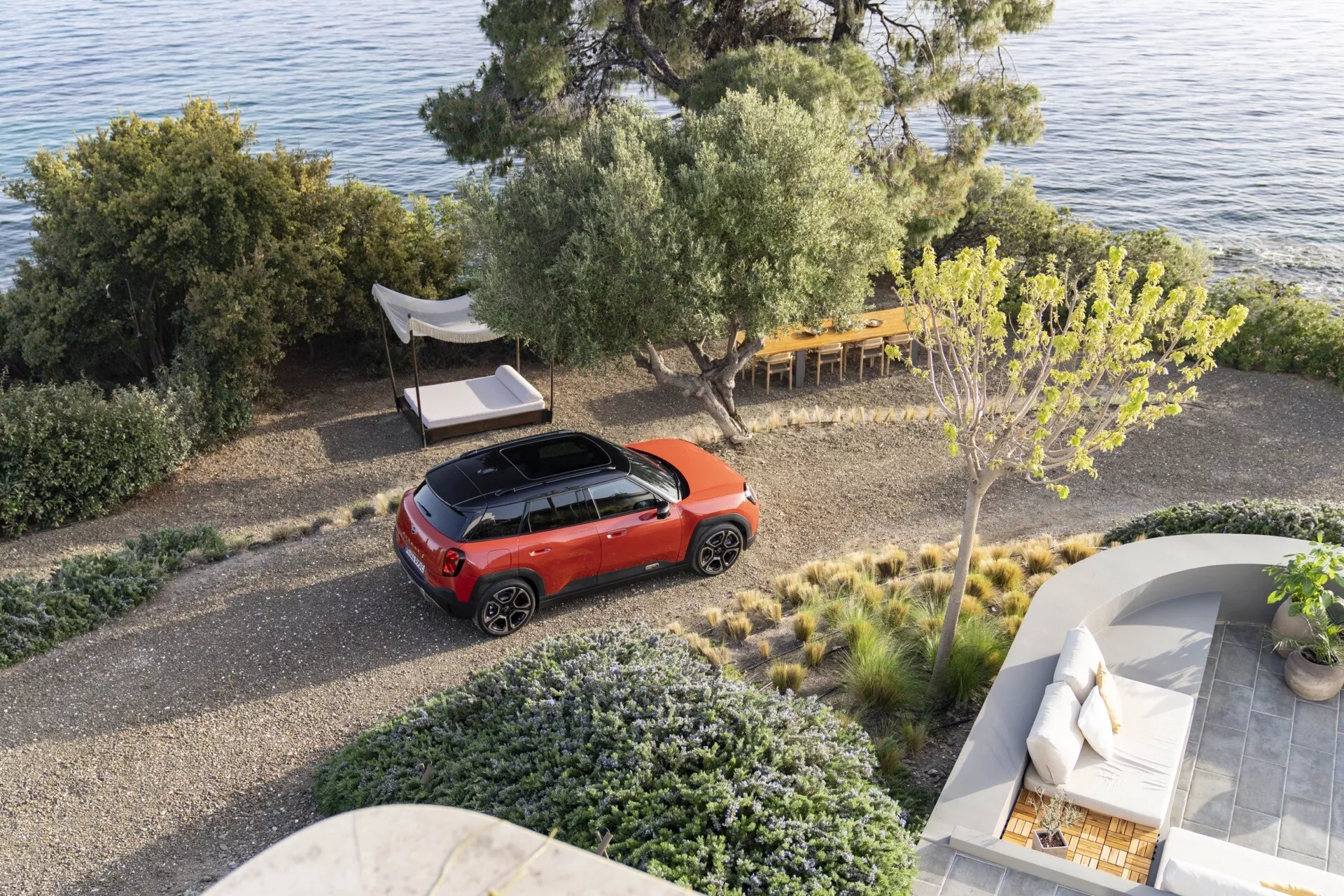 Aerial view of a Mini Aceman 2024 (J05) in bright red with a contrasting dark roof parked on a pebble driveway in a natural setting near a coastline, showcasing its modern crossover design and hinting at advanced automotive technologies.