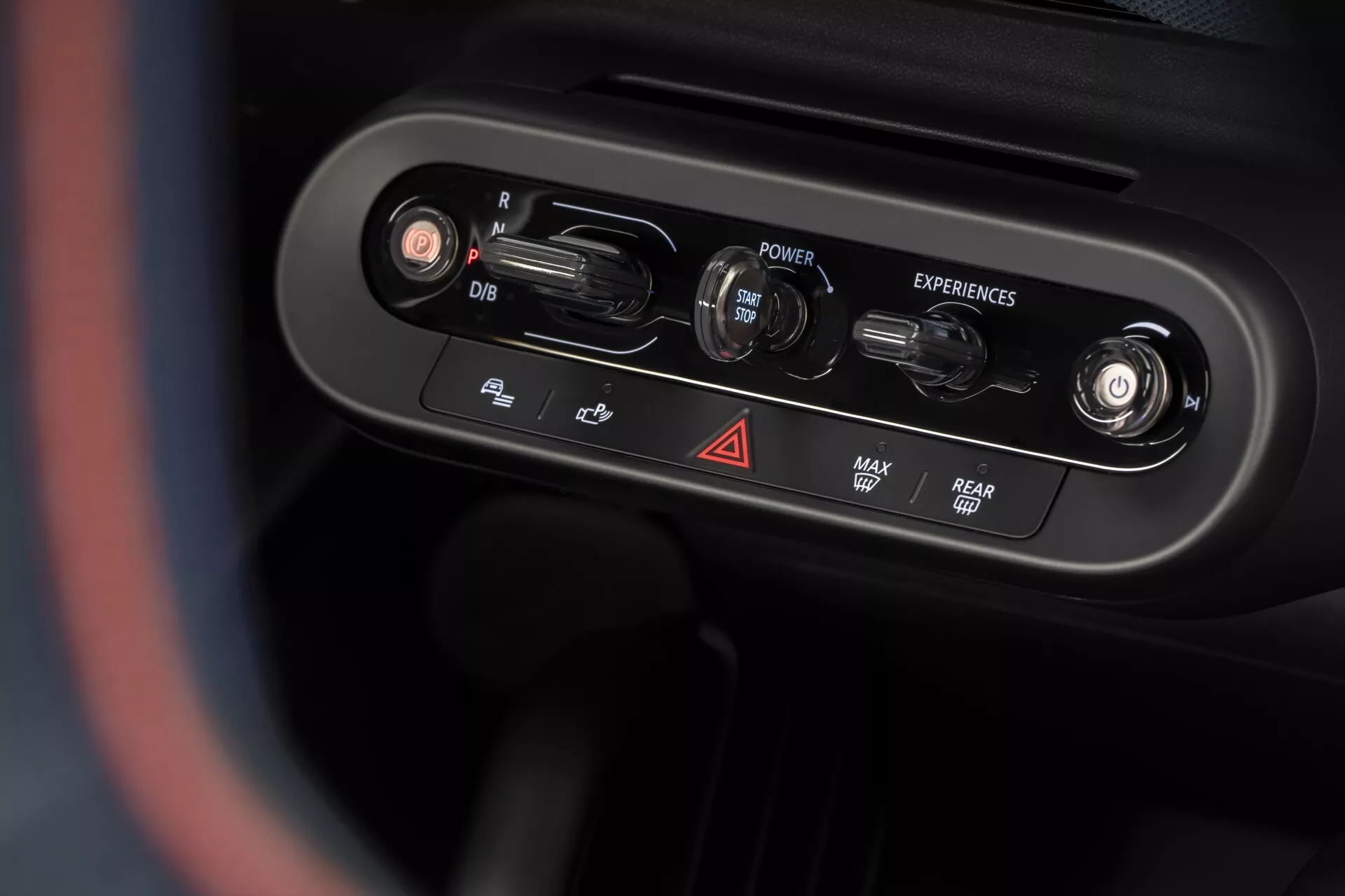 Close-up view of the 2024 Mini Aceman (J05) center console control panel from driver's perspective, featuring rotary dials and buttons for vehicle functions including rearview mirror adjustment, power ignition, drive experiences selection, hazard lights, defrost, rear window heating, and heated steering wheel, with a premium chrome finish