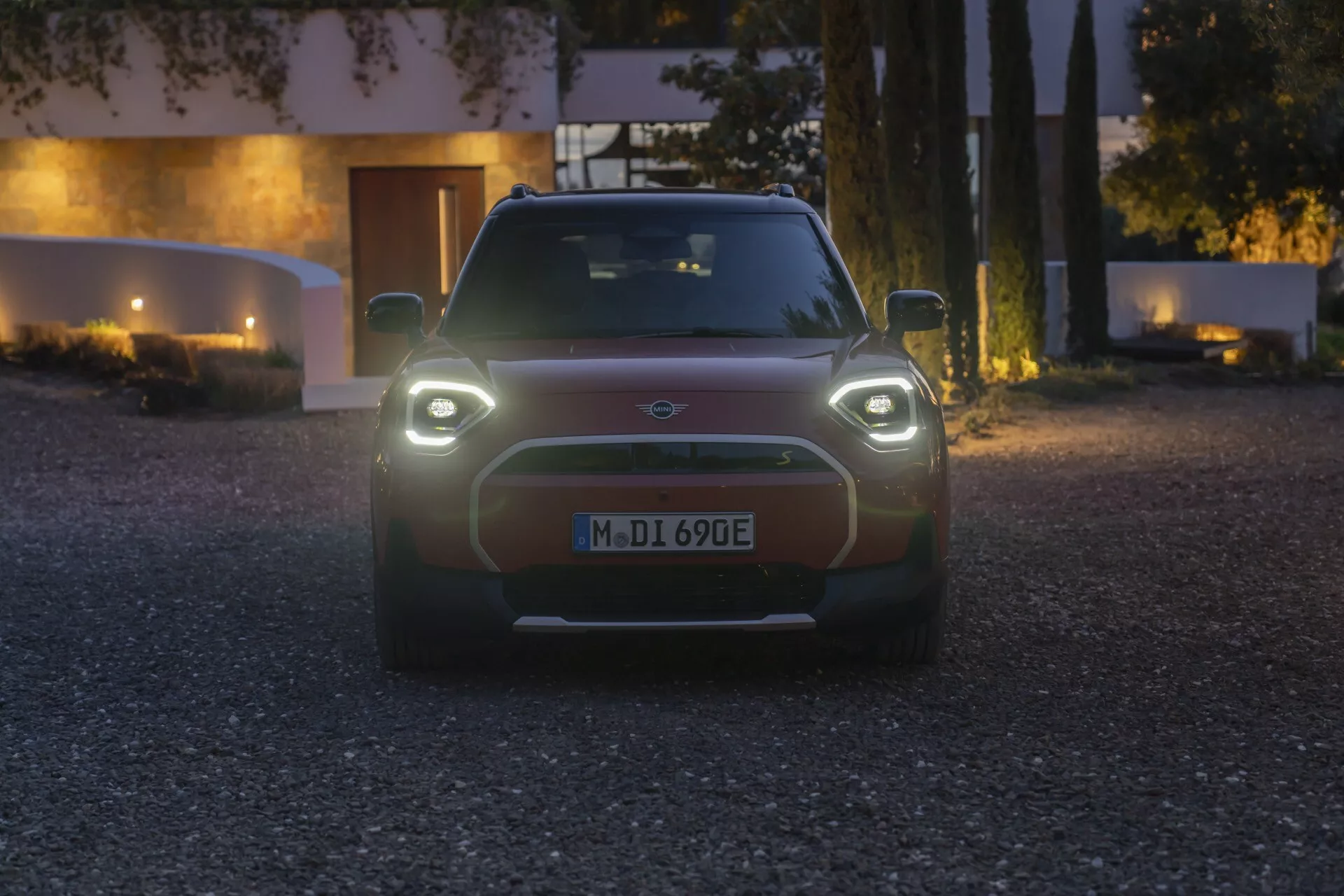 Frontal view of 2024 Mini Aceman (J05) electric car during twilight with illuminated circular headlights and minimalistic grille, showcasing modern EV design