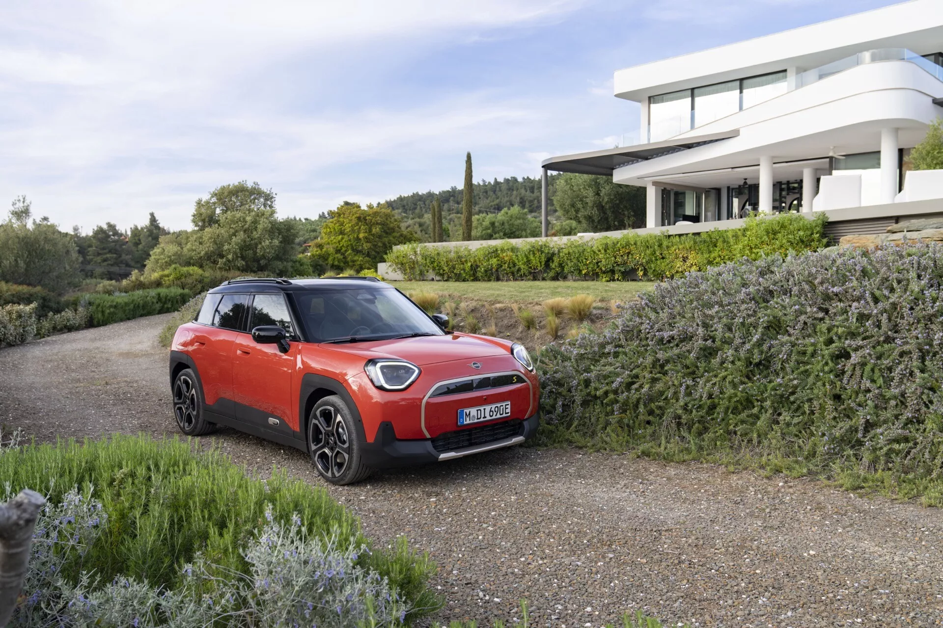 2024 Mini Aceman Subcompact SUV in red with black roof, showcasing modern LED headlights and aerodynamic alloy wheels parked on gravel driveway with greenery and upscale home in background