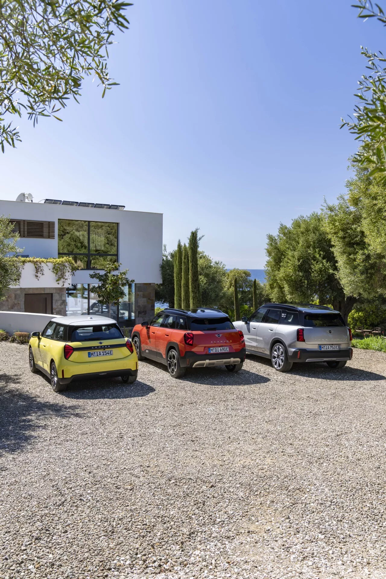 2024 Mini Aceman (J05) triple lineup, showing rear angles with brightly colored finishes of yellow, red, and light color parked side by side on gravel, featuring distinct design elements like wrap-around tail lights, chrome accents, and SUV shapes, with backdrop of residential setting