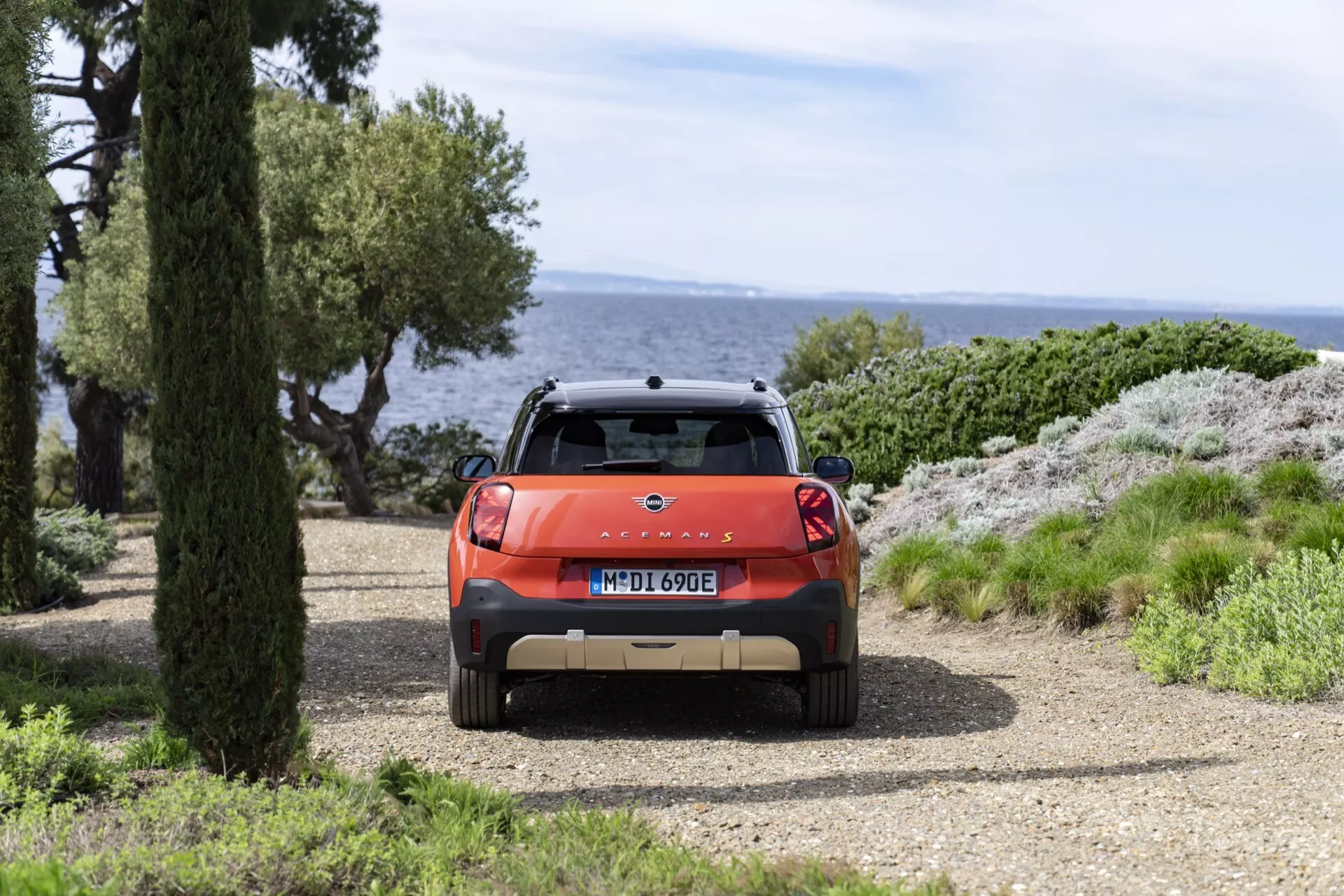 Rear view of the 2024 Mini Aceman (J05) in orange, featuring a sporty spoiler and LED tail lights, positioned on a gravel road by the sea with natural scenery in the background during daytime