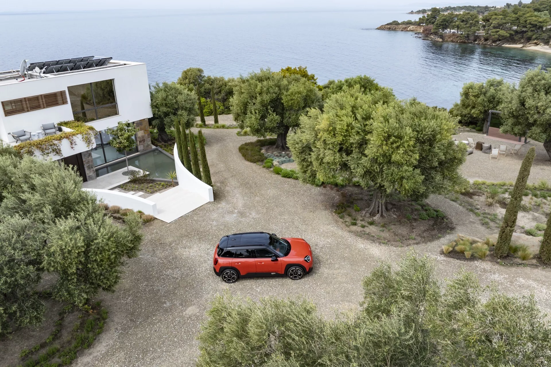 2024 Mini Aceman (J05) red hatchback parked on a gravel driveway with aerial view showcasing the full top side of the vehicle, amidst a scenic landscape with trees, shrubbery, and a sea backdrop next to a modern house