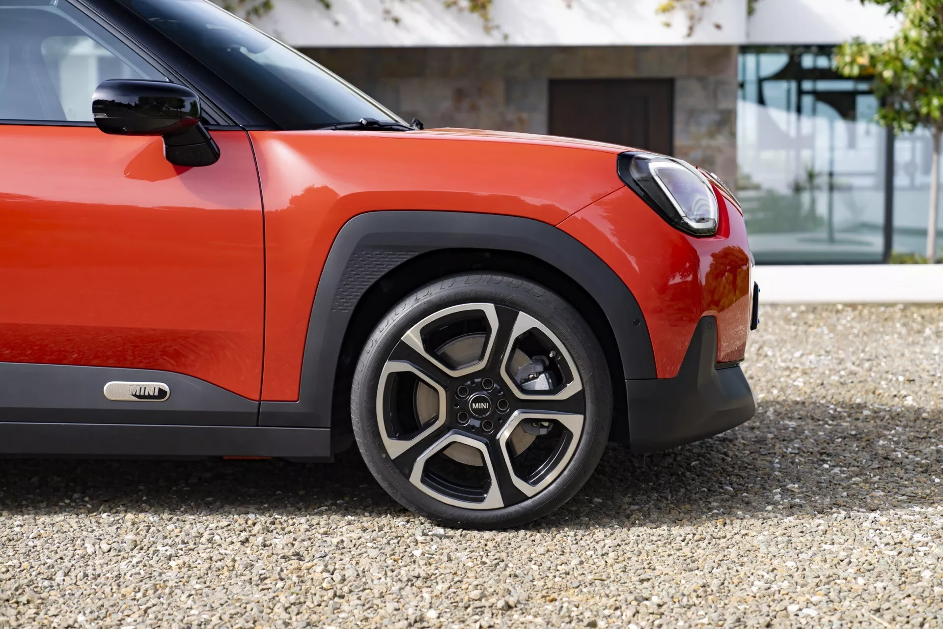 2024 Mini Aceman (J05) showcasing a close-up side view of the sporty front wheel with a distinctive multi-spoke design and two-toned rim, complemented by the front quarter's bright reddish-orange and black two-tone bodywork, and a glimpse of advanced LED headlight technology