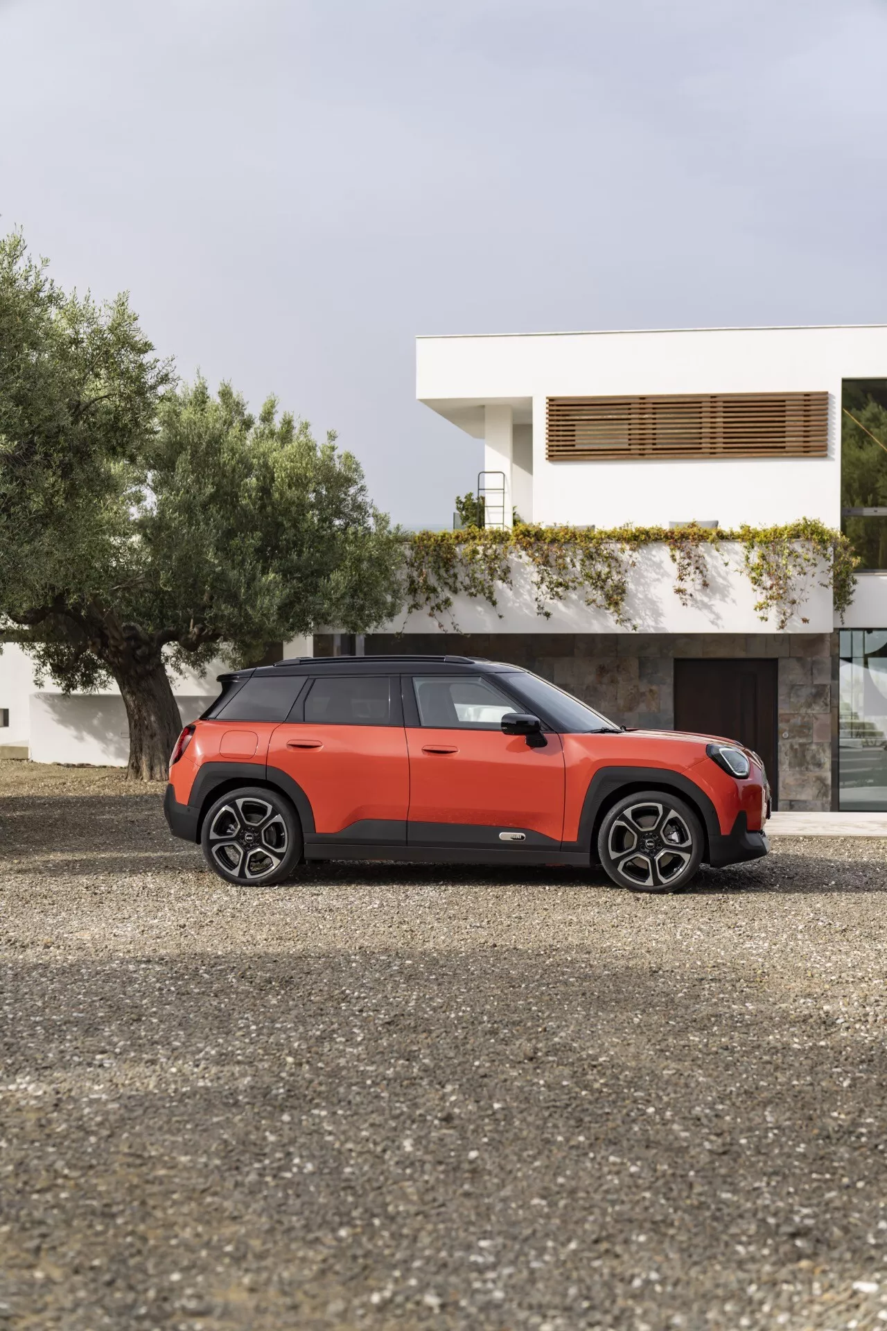 2024 Mini Aceman (J05) parked on gravel outside a modern house, displaying its side profile with a three-quarter view showcasing two-tone color scheme, flush door handles, and multi-spoke wheels