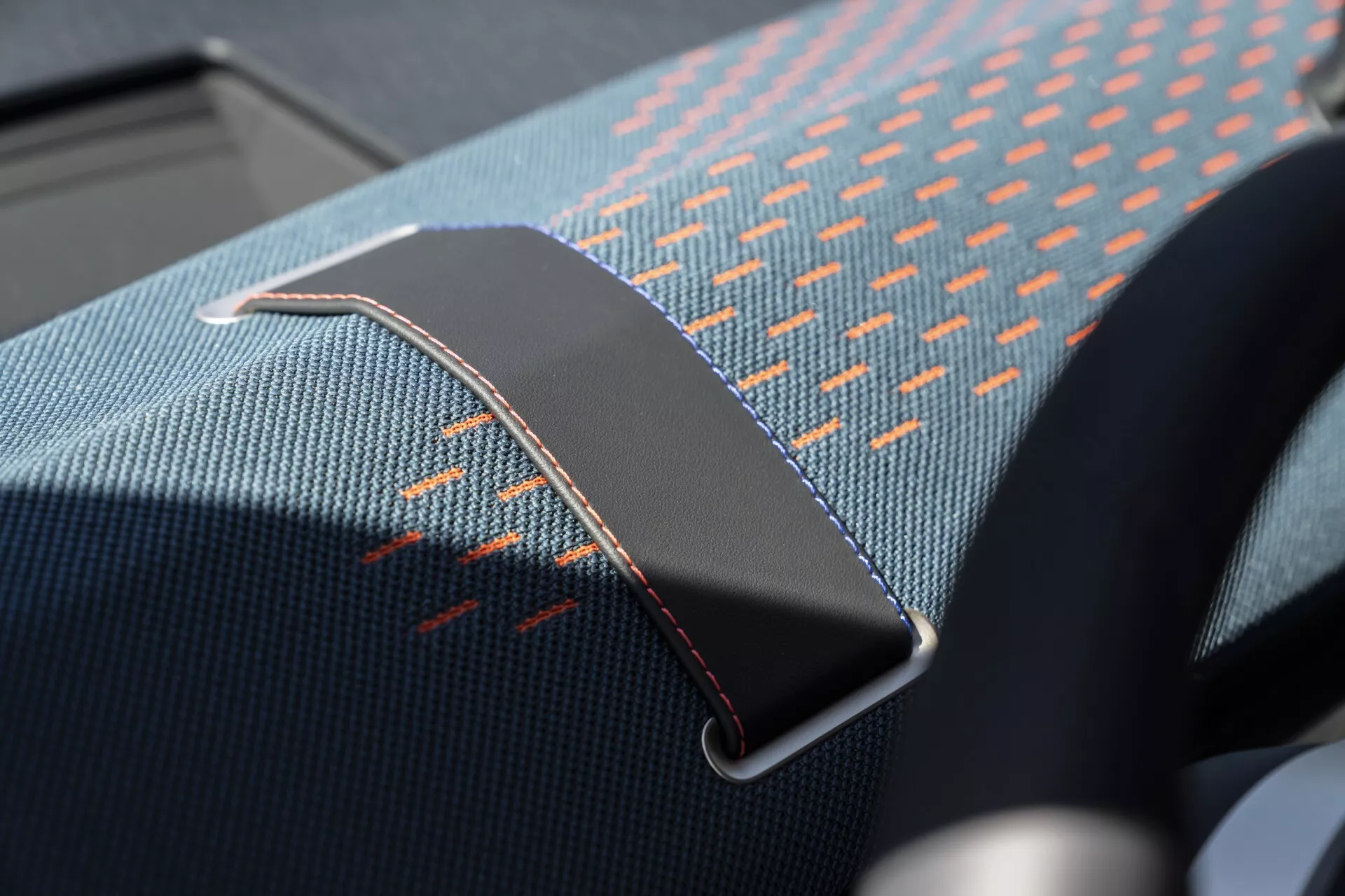 Close-up image of a 2024 Mini Aceman (J05) interior highlighting the seatbelt clip, featuring blue and orange patterned seat fabric with detailed orange stitching and a metallic seatbelt buckle with chromium finish and maroon stitching accents.