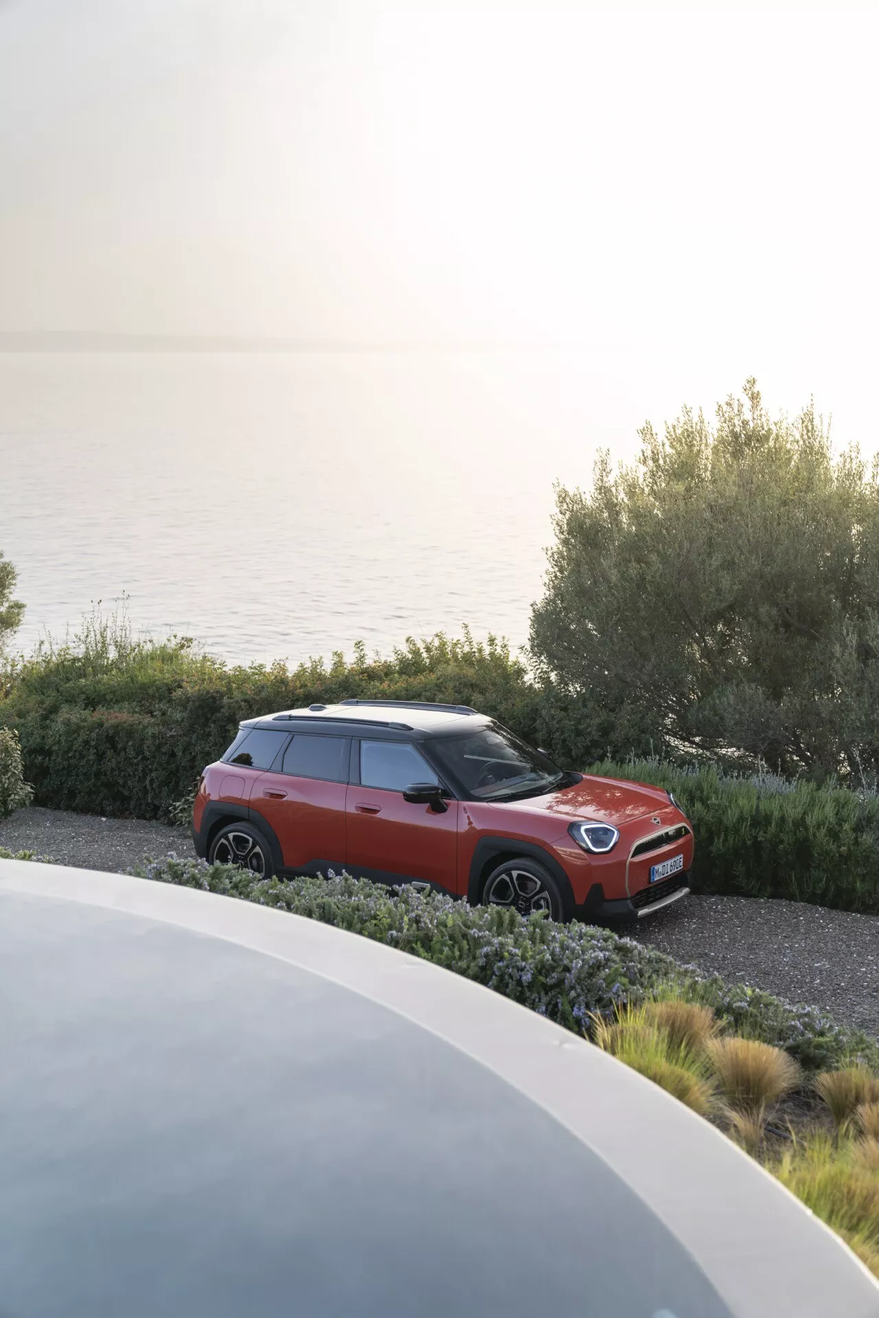 2024 Mini Aceman (J05) parked in a scenic coast with two-tone red and black body, featuring LED headlights, alloy wheels, integrated turn signal mirrors, tinted windows, and roof rails, viewed from an elevated front left quarter angle