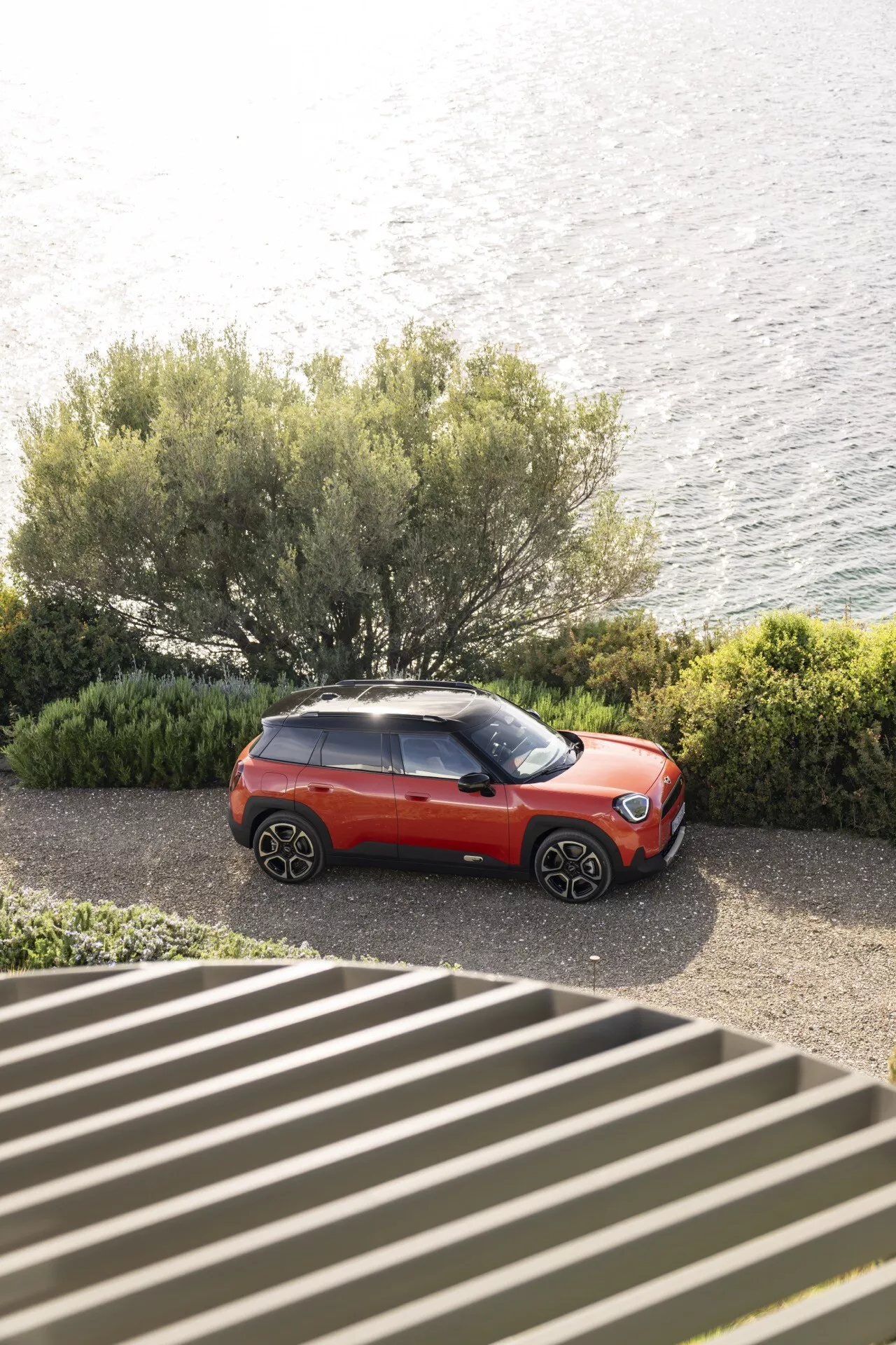 2024 Mini Aceman (J05) in red and black, viewed from above, showcasing stylish alloy wheels, modern LED headlights, and two-tone paint job, parked by the shoreline