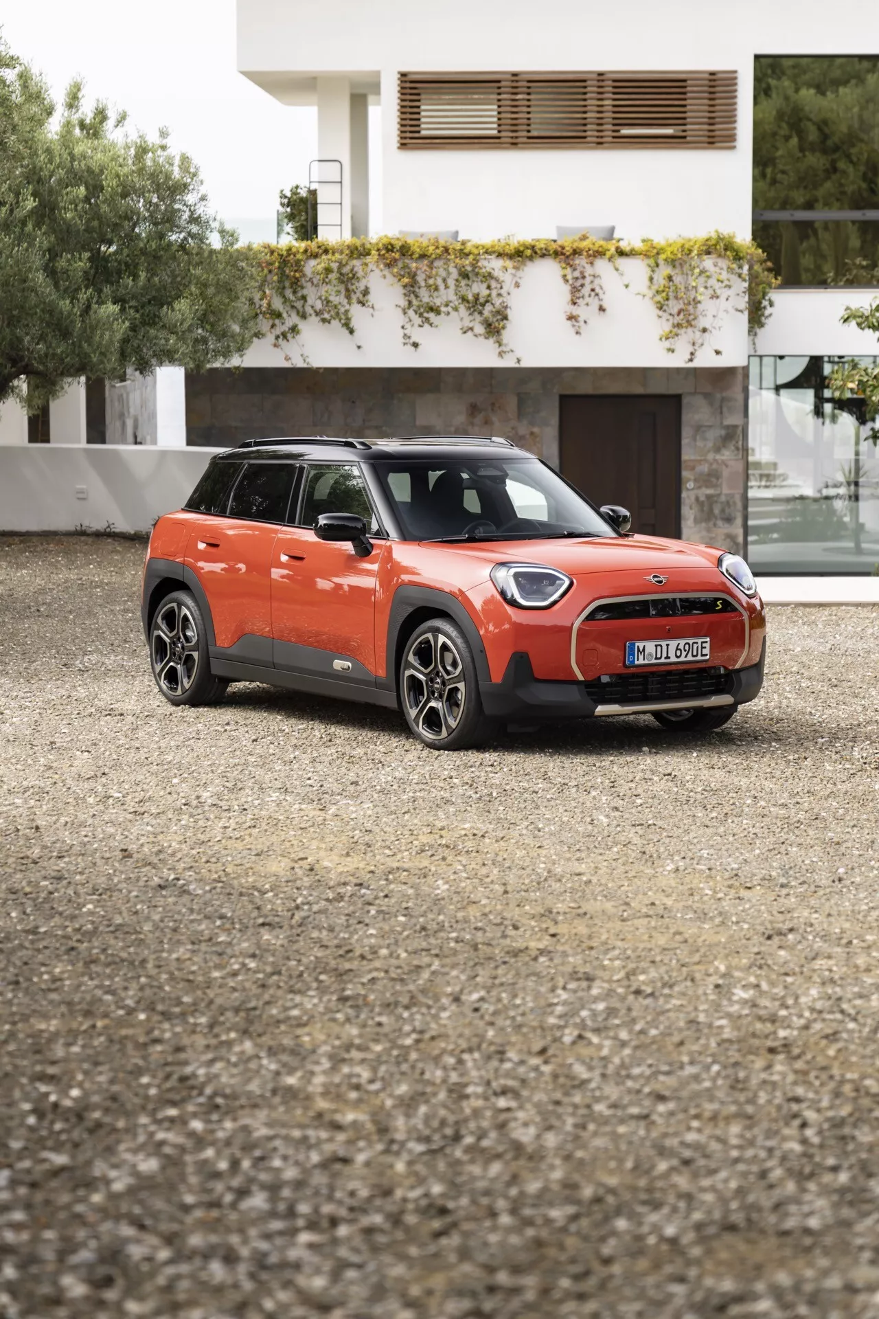 2024 Mini Aceman (J05) compact SUV in vibrant orange with black roof, displayed from a front-quarter angle on a gravel surface, featuring modern LED headlights, alloy wheels, and an aerodynamic grille design against a residential backdrop.