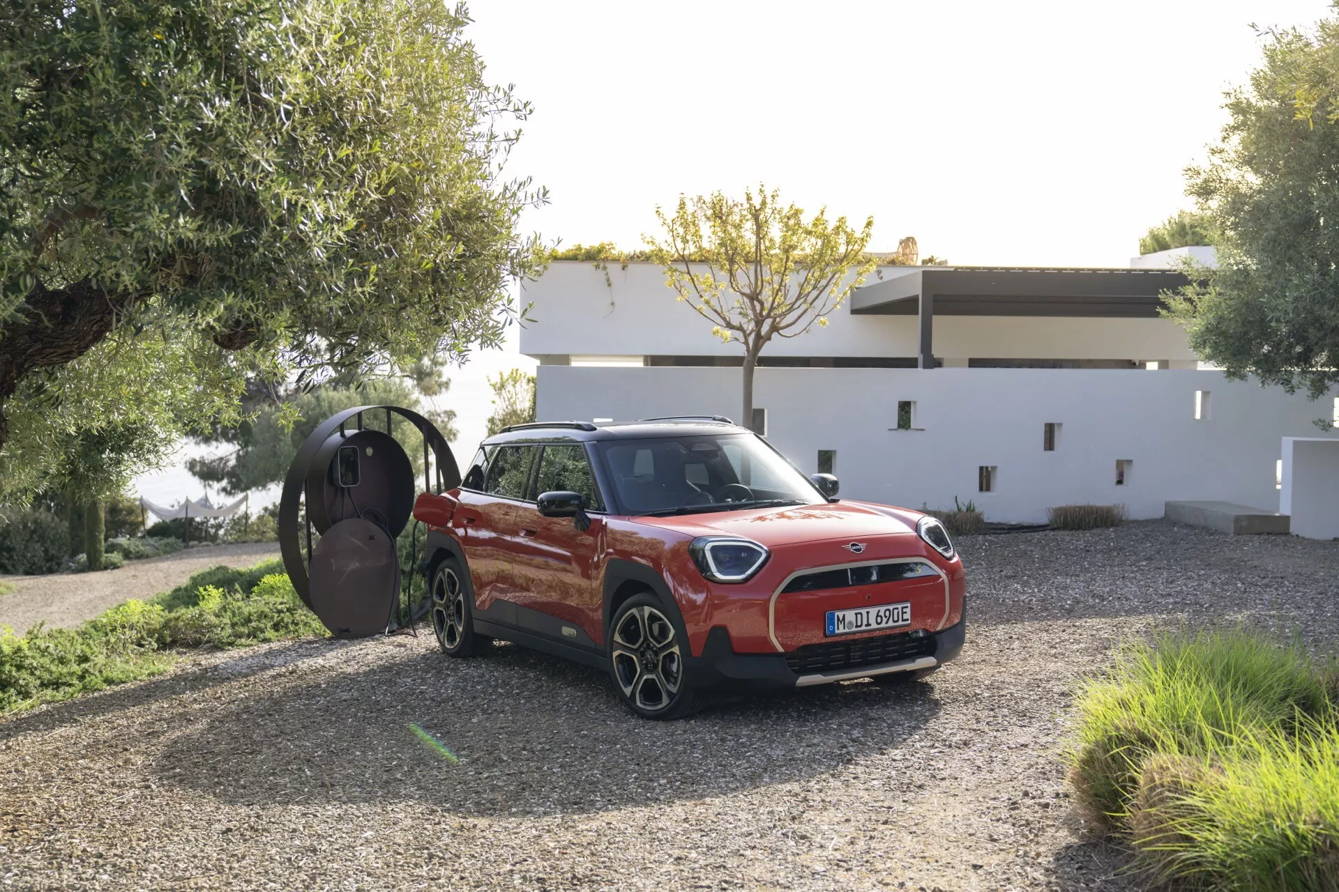2024 Mini Aceman (J05) in vibrant red displayed in a three-quarter frontal view, highlighting its grille, headlights, and open driver-side door, parked on a gravel driveway against a residential backdrop