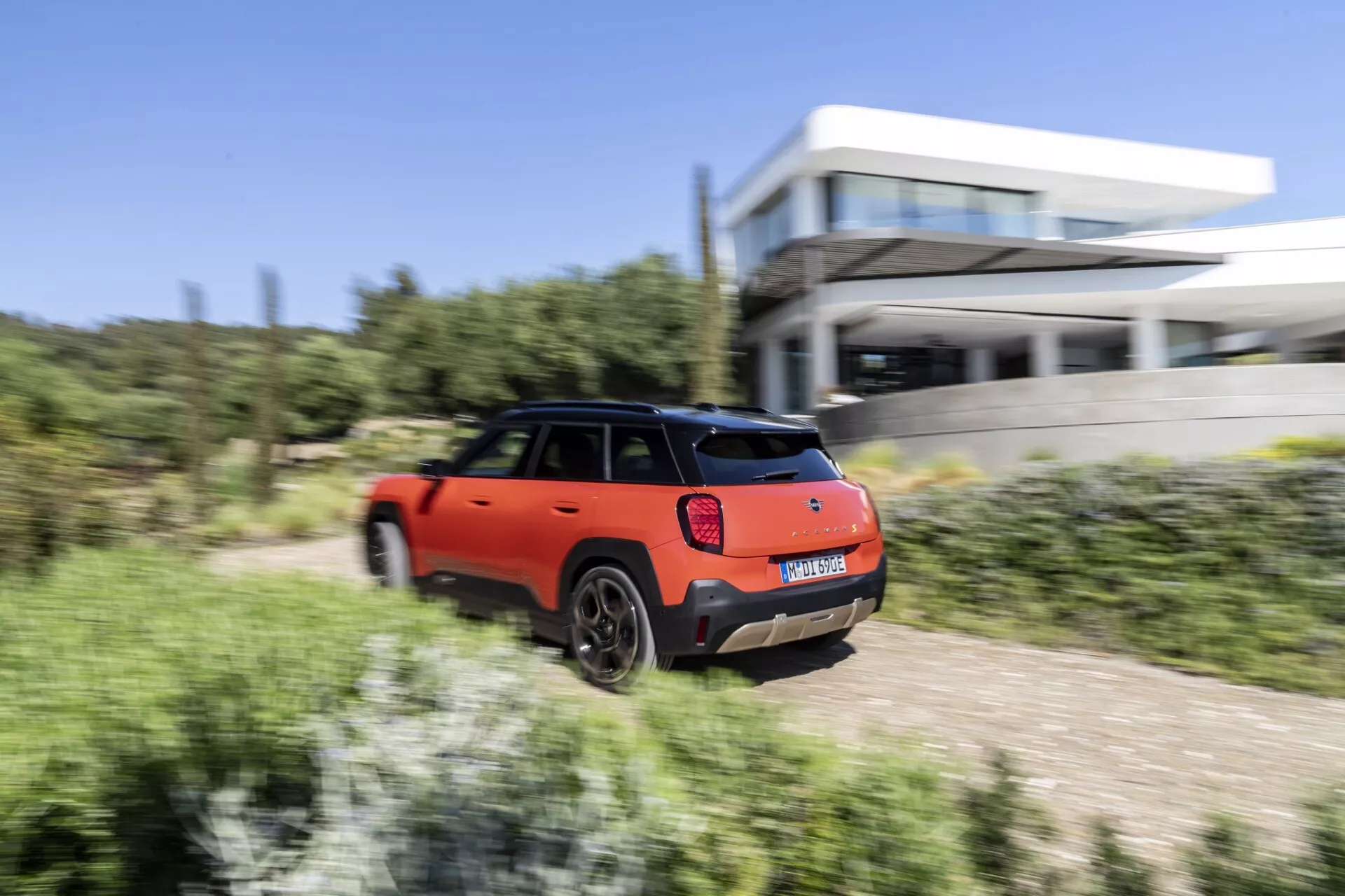 2024 Mini Aceman (J05) in motion, rear three-quarter view highlighting its orange and black two-tone paint, LED tail lights, and sporty aesthetics against a dynamic backdrop featuring natural vegetation and modern architecture