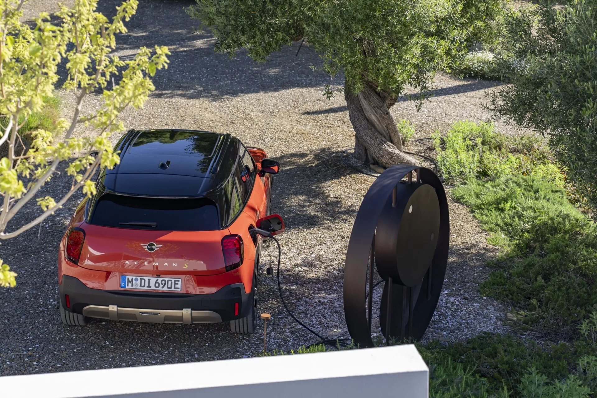 High-angle rear view of a 2024 Mini Aceman (J05) red hatchback electric vehicle connected to a modern charging station, parked outdoors surrounded by trees and gravel.