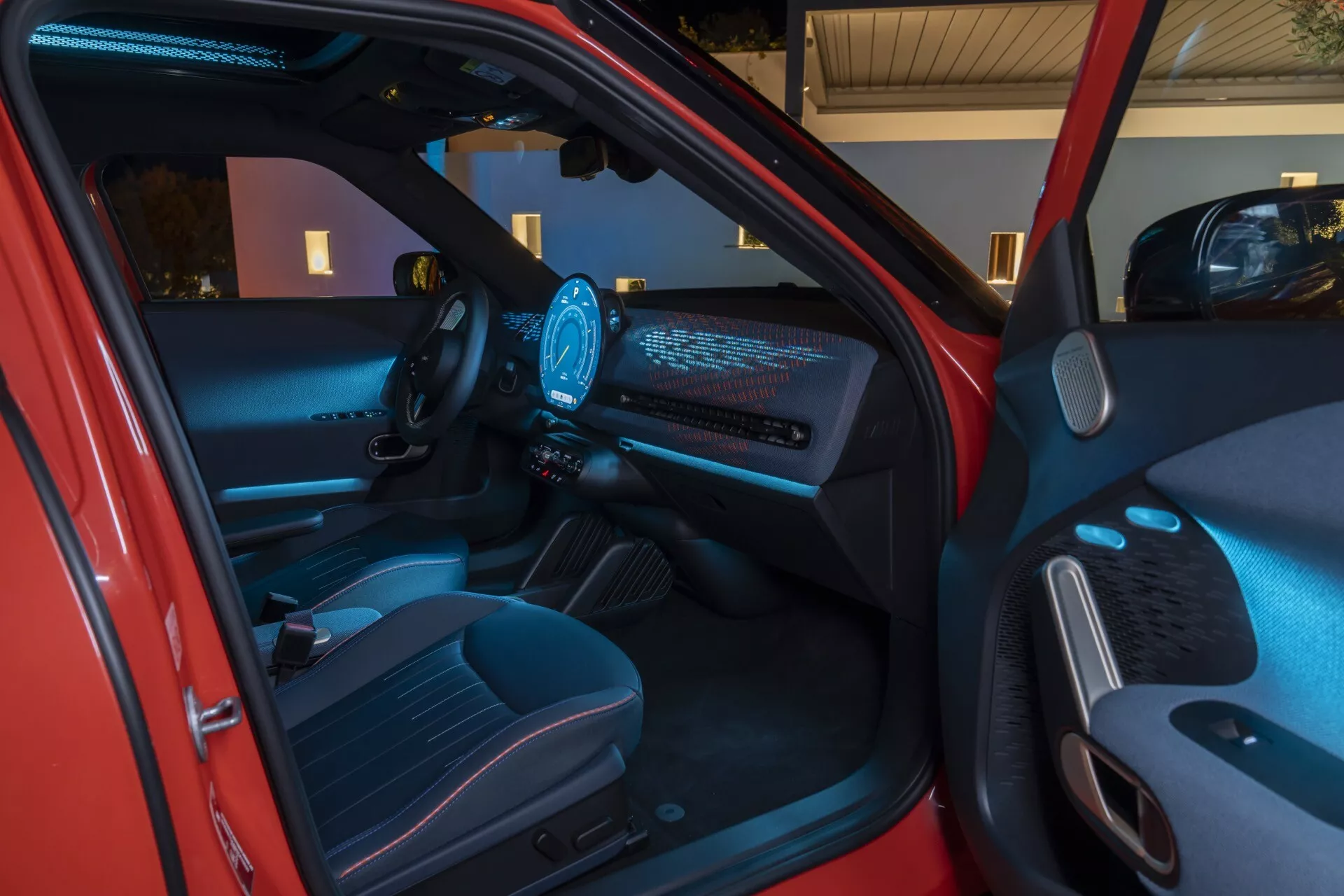 Interior view of the 2024 Mini Aceman (J05) showcasing futuristic dashboard with illuminated display, modern steering wheel with illuminated controls, digital instrument cluster, and high-tech ambient lighting from an exterior angle near the driver's side.