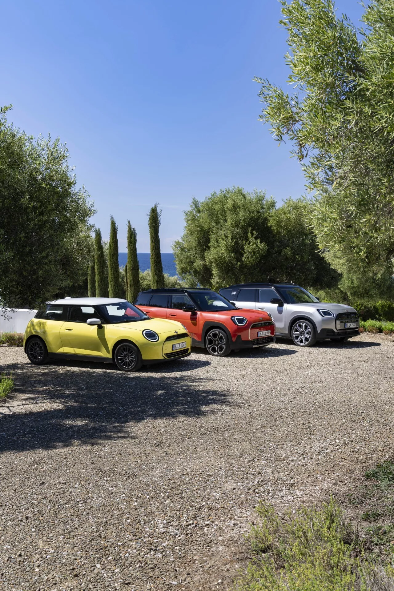 2024 Mini Aceman lineup featuring a bright yellow compact hatchback, vibrant red compact model, and a light metallic SUV parked side by side on gravel with green foliage and clear blue sky in the background, highlighting modern designs and technological features