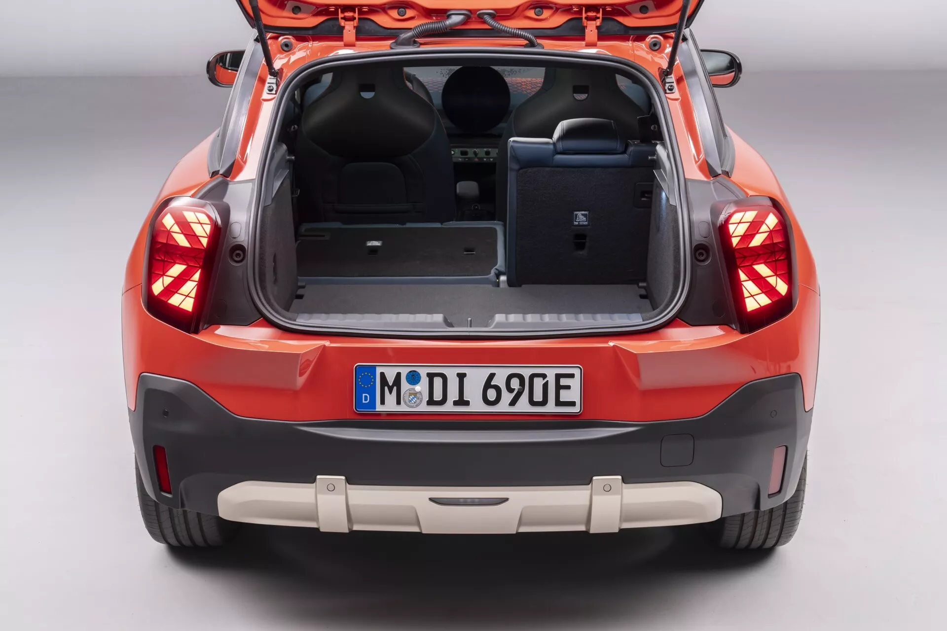 Rear view of the 2024 Mini Aceman (J05) highlighting its spacious trunk with an open tailgate, interior trim details, integrated parking sensors on the bumper, and advanced rear lighting assembly, parked in a showroom setting