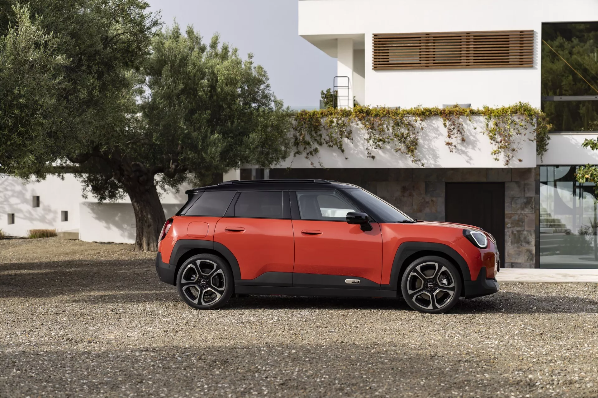 2024 Mini Aceman compact SUV in two-tone red and black parked outdoors in front of a modern residential building, showcasing its side profile with detailed vehicle features and design elements, including multi-spoke alloy wheels, panoramic sunroof, and advanced safety technologies.