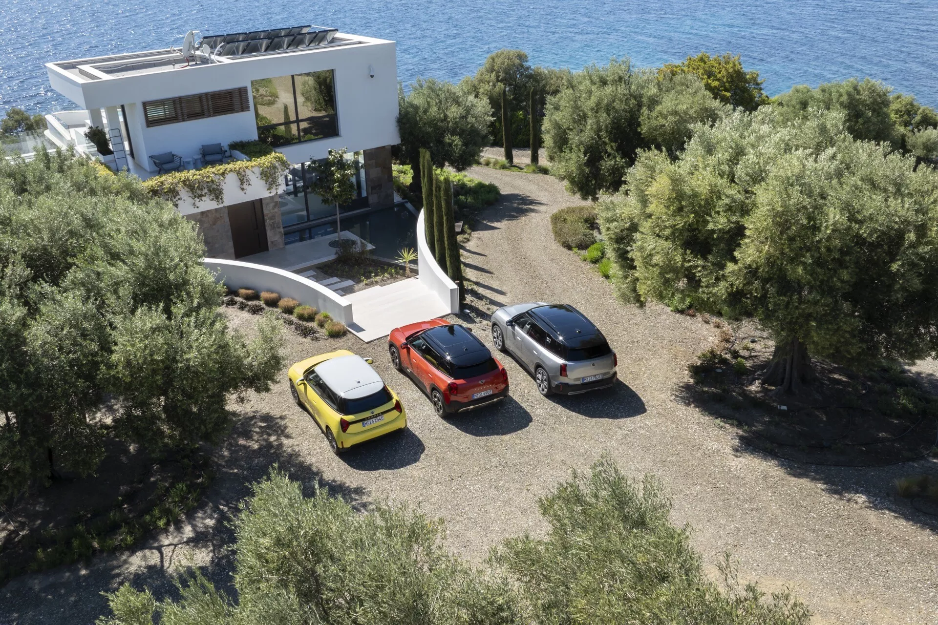 Aerial view of a yellow hatchback, metallic grey sedan, and red crossover parked outside a contemporary house amidst Mediterranean vegetation
