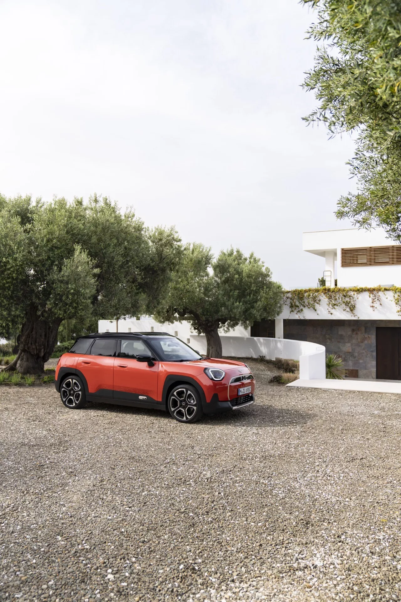 2024 Mini Aceman (J05) in a three-quarter front view highlighting its sleek front grille, LED headlights, and partial side profile with driver's side door and windows on a gravel surface, showcasing modern urban design elements and potential advanced technological features like alloy wheels and a panoramic sunroof against a residential backdrop.