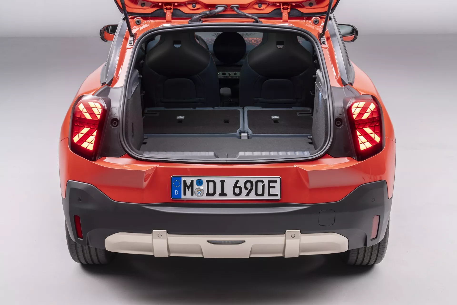Rear view of an orange-red Mini Aceman 2024 (J05) with open hatchback, showcasing empty cargo space, advanced LED taillights, parking assistance sensors, rearview camera, and potential electric vehicle indication from the European-style license plate, against a studio background