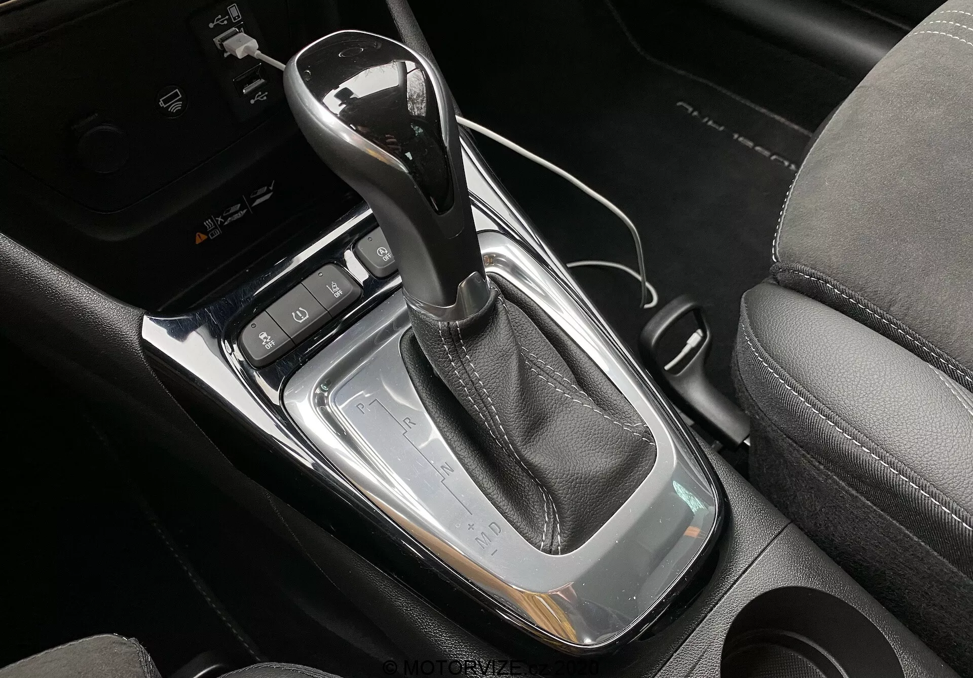 Close-up view of the center console in a modern Opel Crossland 2021, highlighting the automatic gear shifter wrapped in black leather with chrome trim, set into a glossy black panel with silver borders. The image also shows a black button with a P for electronic parking brake, two cup holders, and dynamic control buttons near the gear shifter, with a partially visible black leather seat.
