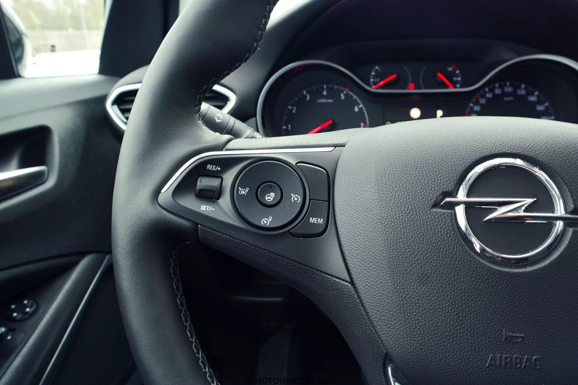 Close-up view of a premium car's steering wheel and driver's cockpit, featuring a prominently displayed brand logo with a circle and lightning bolt. The leather-textured steering wheel is equipped with multifunctional control buttons for cruise control, volume adjustment, and multimedia navigation, suggesting a focus on comfort and advanced technology. The partially visible instrument cluster combines analog gauges with red needles, underlining a blend of analog and digital elements. The shallow depth of field highlights the steering controls, maintaining viewer's focus on ergonomic design and control accessibility from a driver's perspective, angled slightly to the left.