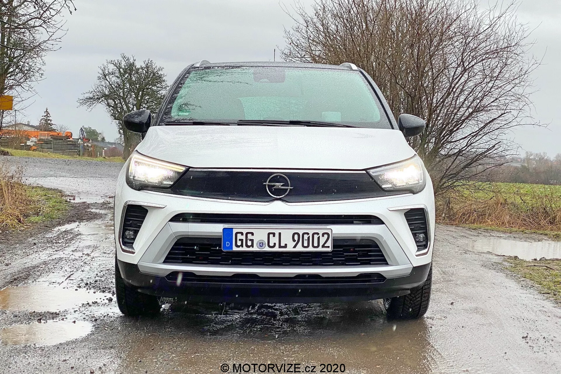 Front angle view of a 2021 Opel Crossland showcasing detailed features including the Opel badge on the grille, sleek LED daytime running headlights, two-tone white and black color scheme, front bumper with fog lights, wide air intakes, and a noticeable front-facing camera indicating advanced driver-assist technologies. The car is stationary on wet asphalt with visible water droplets on the vehicle, emphasizing weather-related driving conditions.