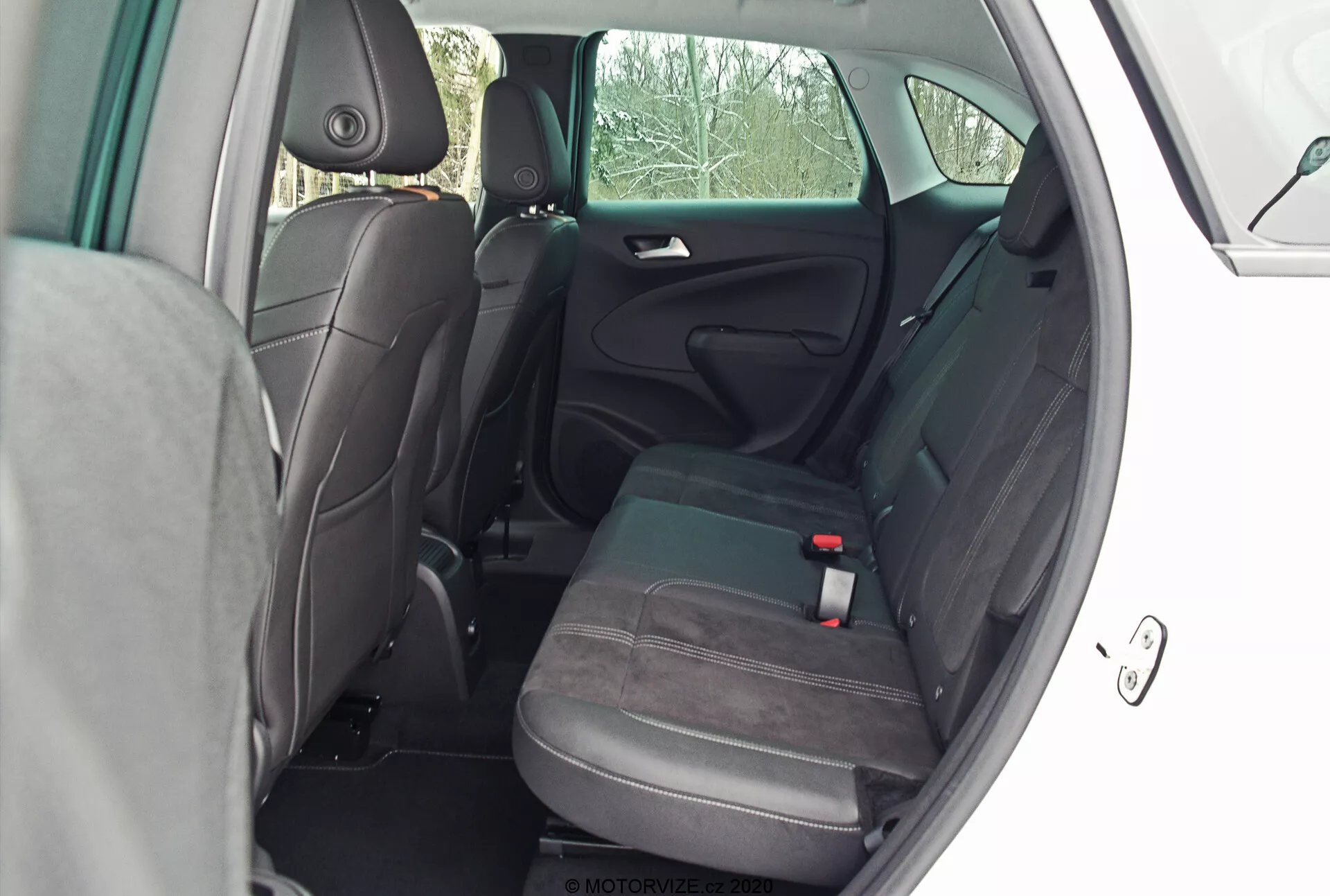 Rear passenger compartment of the 2021 Opel Crossland, viewed from the external side angle with the rear door open. The photo, taken from a perspective slightly behind the rear door, offers a clear view into the cabin, showcasing the two-tone fabric upholstery in grey and black, adjustable headrests, three-point seatbelt for the middle seat, ISOFIX child car seat anchor, substantial legroom, center armrest, storage pocket on the front passenger seat's back, and the door panel with a door handle, speaker grille, and trim.