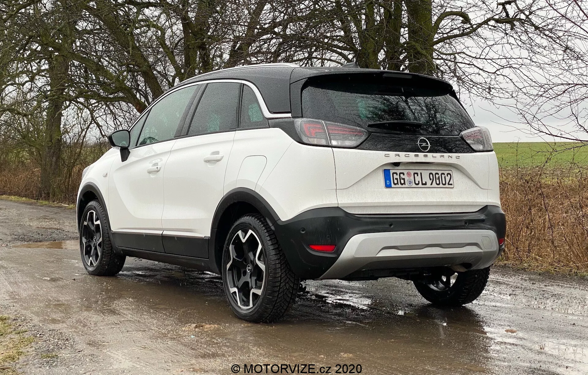 Modern compact SUV Opel Crossland 2021 in a three-quarter rear view angle from rear-left perspective, displaying white color with black accents, contrasting black roof and side mirrors, black cladding along the lower edges and wheel arches, horizontally oriented LED tail lights, manufacturer's badge on the tailgate, parking sensors in the rear bumper, styled alloy wheels with dark finish, parked on a country road with wet surfaces suggesting recent rain or moist environment.