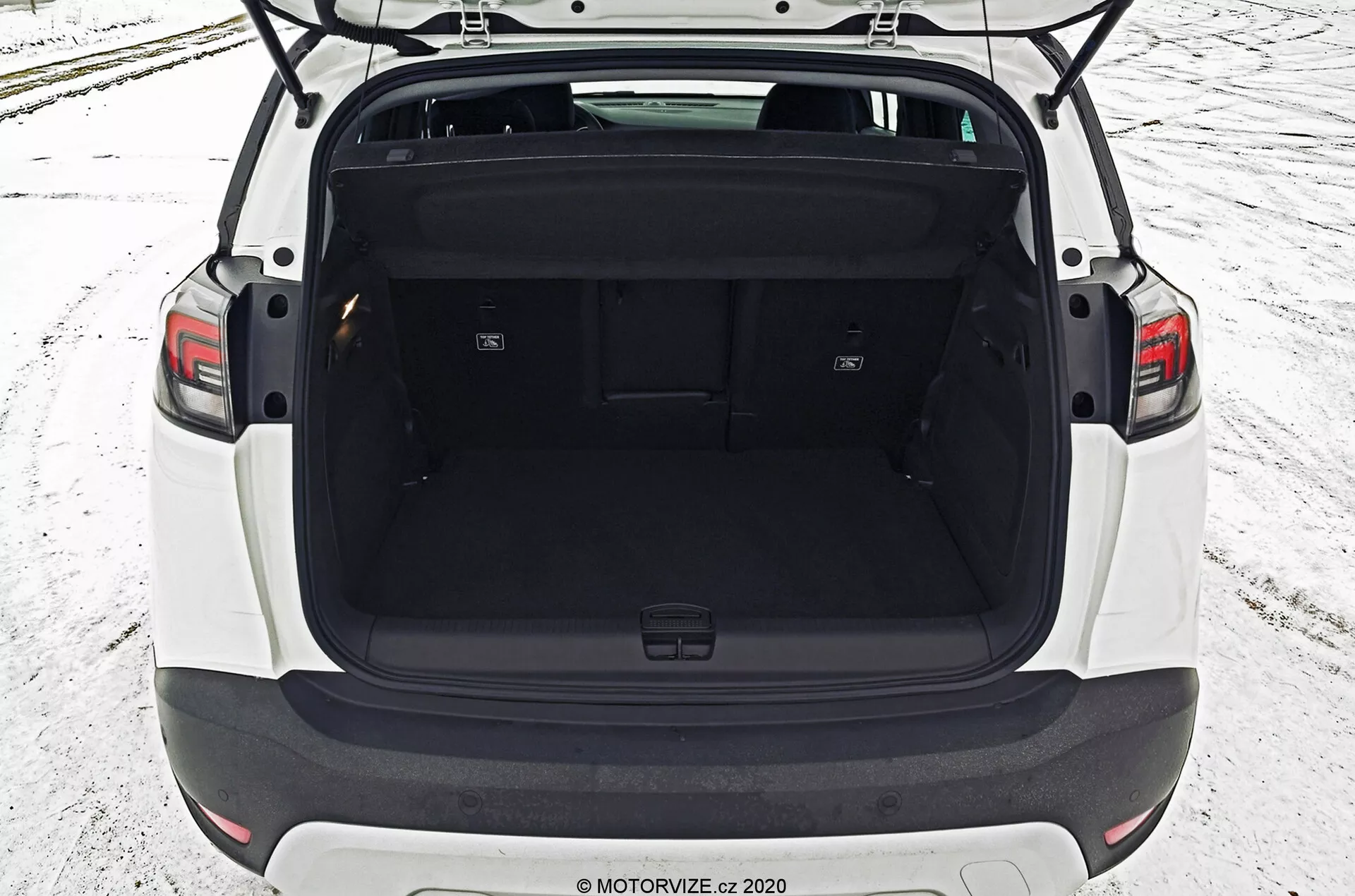 The image displays the rear section of an Opel Crossland 2021, showcasing an elevated and centered perspective of the open trunk space. The rear seats are folded down to create a flat, expanded load area, furnished with accessory hooks and side compartments. The trunk is immaculately clean, suggesting a new or well-maintained vehicle, with dark interior trim and carpeting. No advanced cargo management technology or personal items are visible, underlining the functional and capacious design of this modern hatchback or SUV.