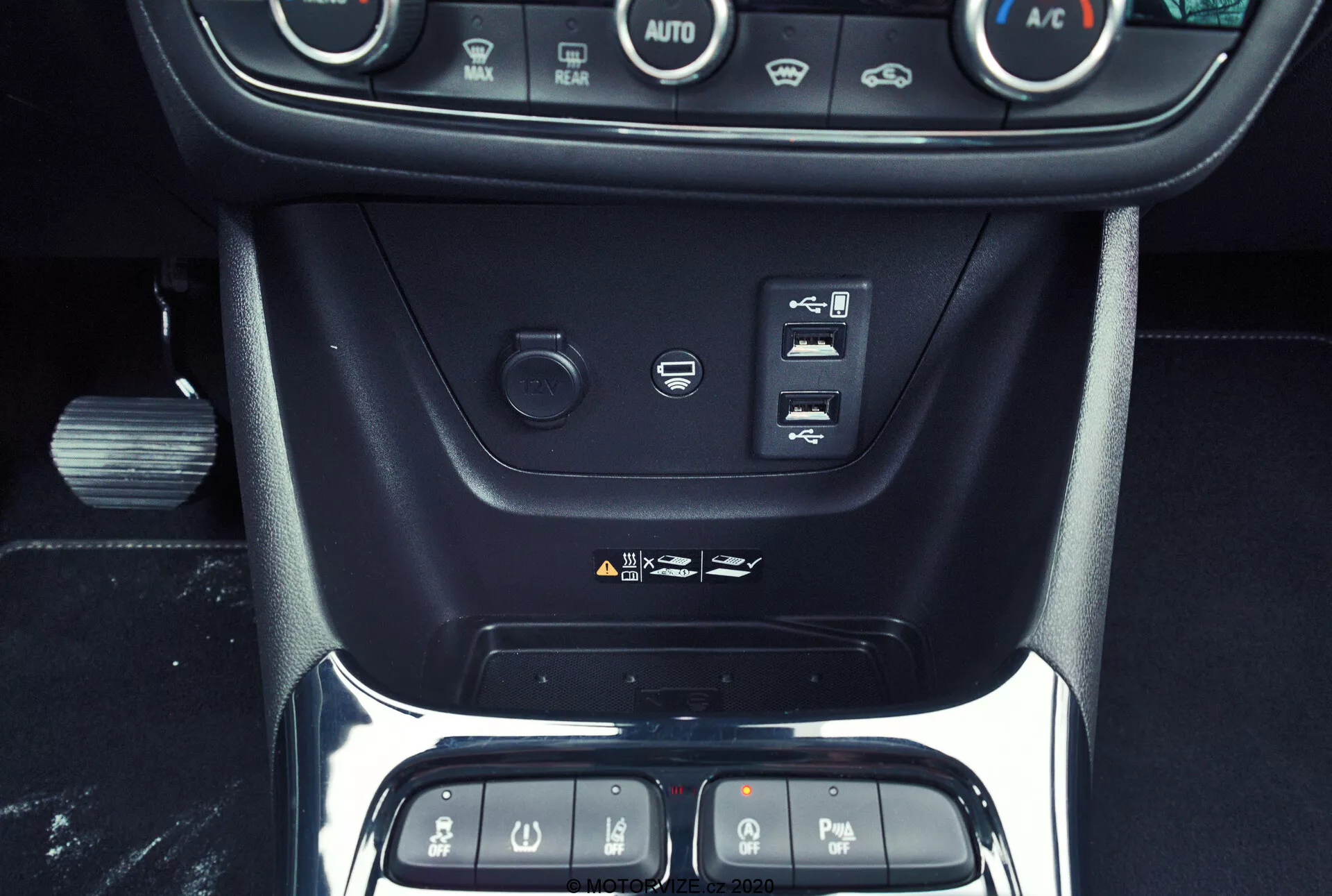 Interior shot of the 2021 Opel Crossland focusing on the center console, showcasing climate control buttons, start/stop engine button, driving assistance buttons, a 12-volt power outlet, USB port, and part of the gear shift area with a chrome-framed glossy black panel, emphasizing the car's modern and premium interior technology and design.