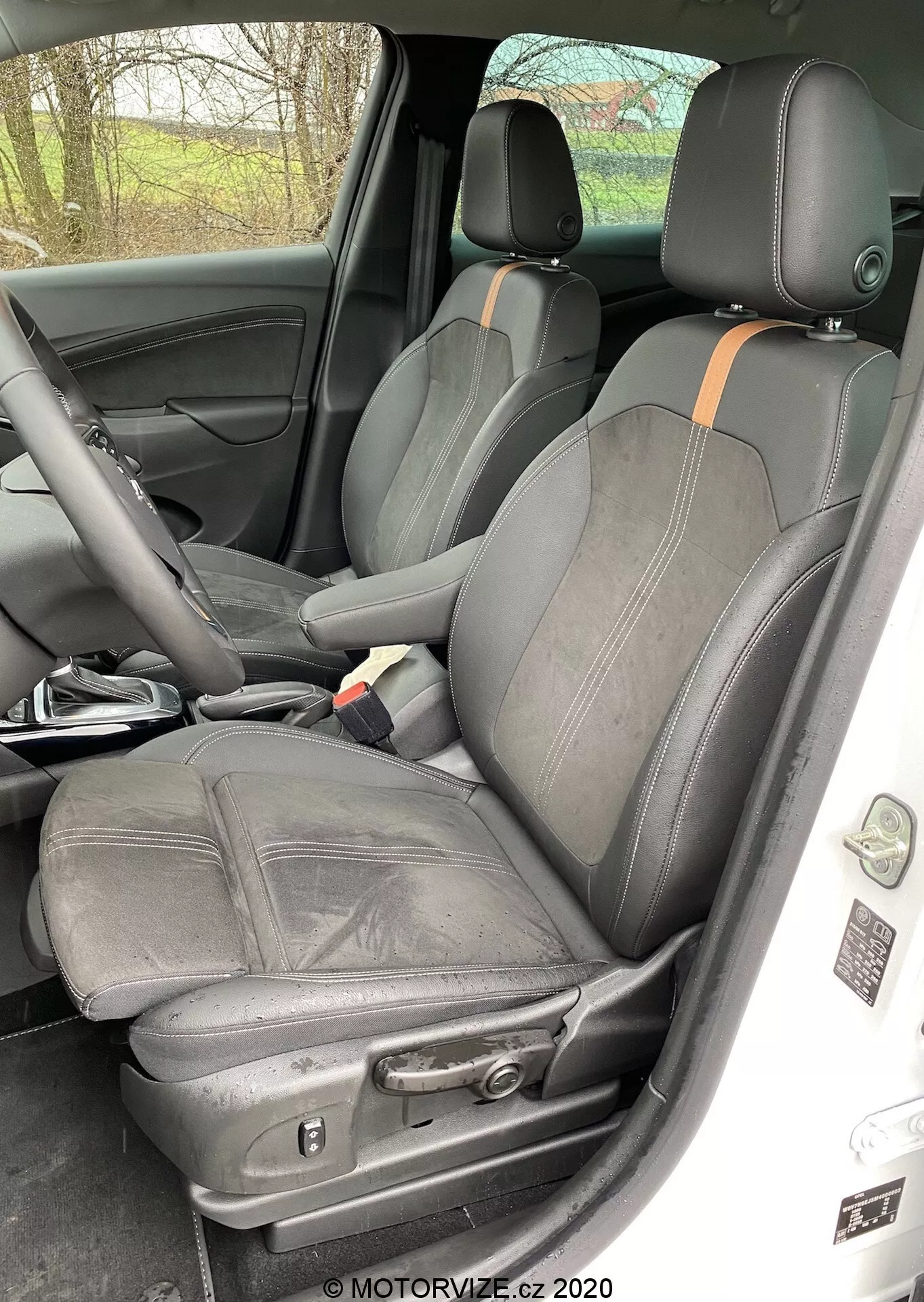 Interior of a 2021 Opel Crossland focusing on front driver and passenger seats, taken from outside the passenger side with open doors. The seats made of black leather and fabric feature detailed stitching and side bolsters for lateral support, with an orange or light brown accent. The driver's seat shows creases, suggesting use, and has electronic adjustment controls. The center console hints at additional features. A speaker grille is visible on the door panel, and a seat belt latch rests on the central armrest, indicating quality materials and construction.