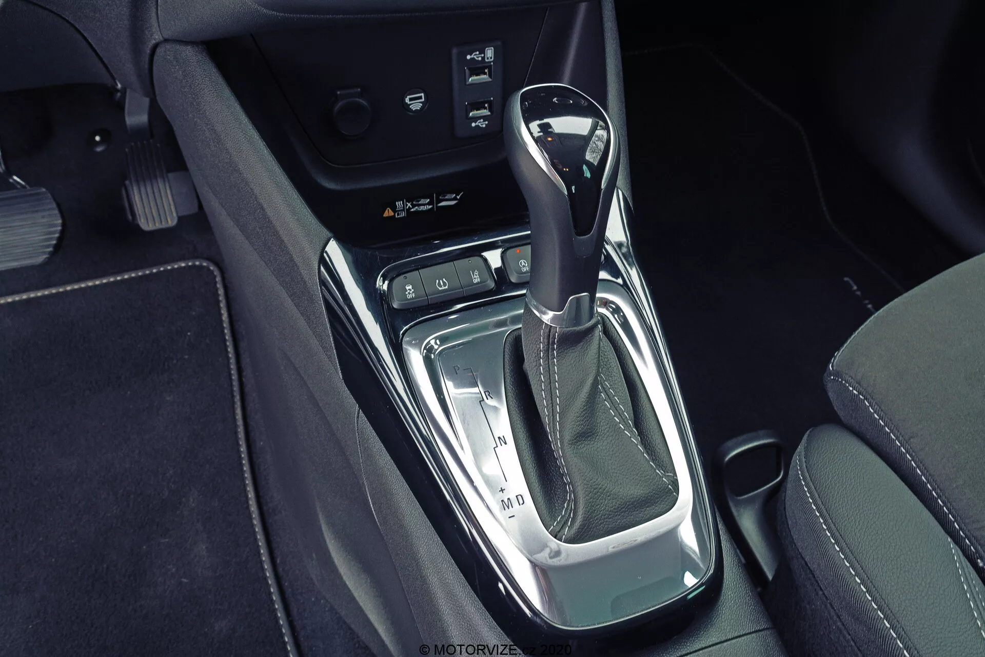 Close-up top-down view of the Opel Crossland 2021 center console featuring an automatic gear shifter with leather-wrapped knob, chrome and piano black finishes, electronic parking brake switch, and possible storage compartment or connectivity panel surrounded by black leather upholstery with contrasting stitching.