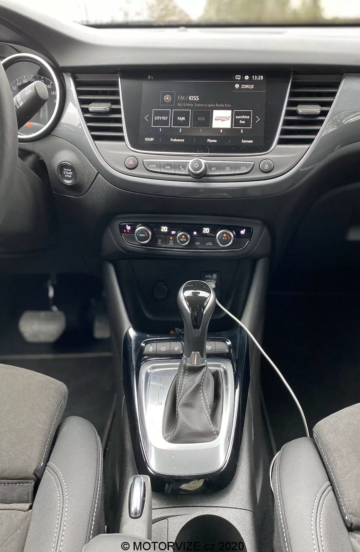 Interior of Opel Crossland 2021 from first-person perspective, showing center console and dashboard. Features modern automatic transmission shifter with leather boot, dual-zone climate control panel, infotainment system with a touch screen, cup holders, and steering wheel with mounted controls.