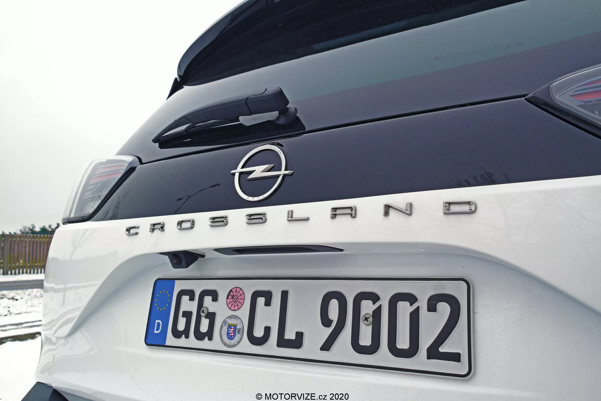 Close-up of the rear part of a 2021 Opel Crossland from the right side at an angled upward perspective, showcasing the trunk, right taillight, rear bumper, and prominent make and model badging with the Opel logo and 'Crossland' text. European license plate and traces of snow, indicating an outdoor, overcast setting.