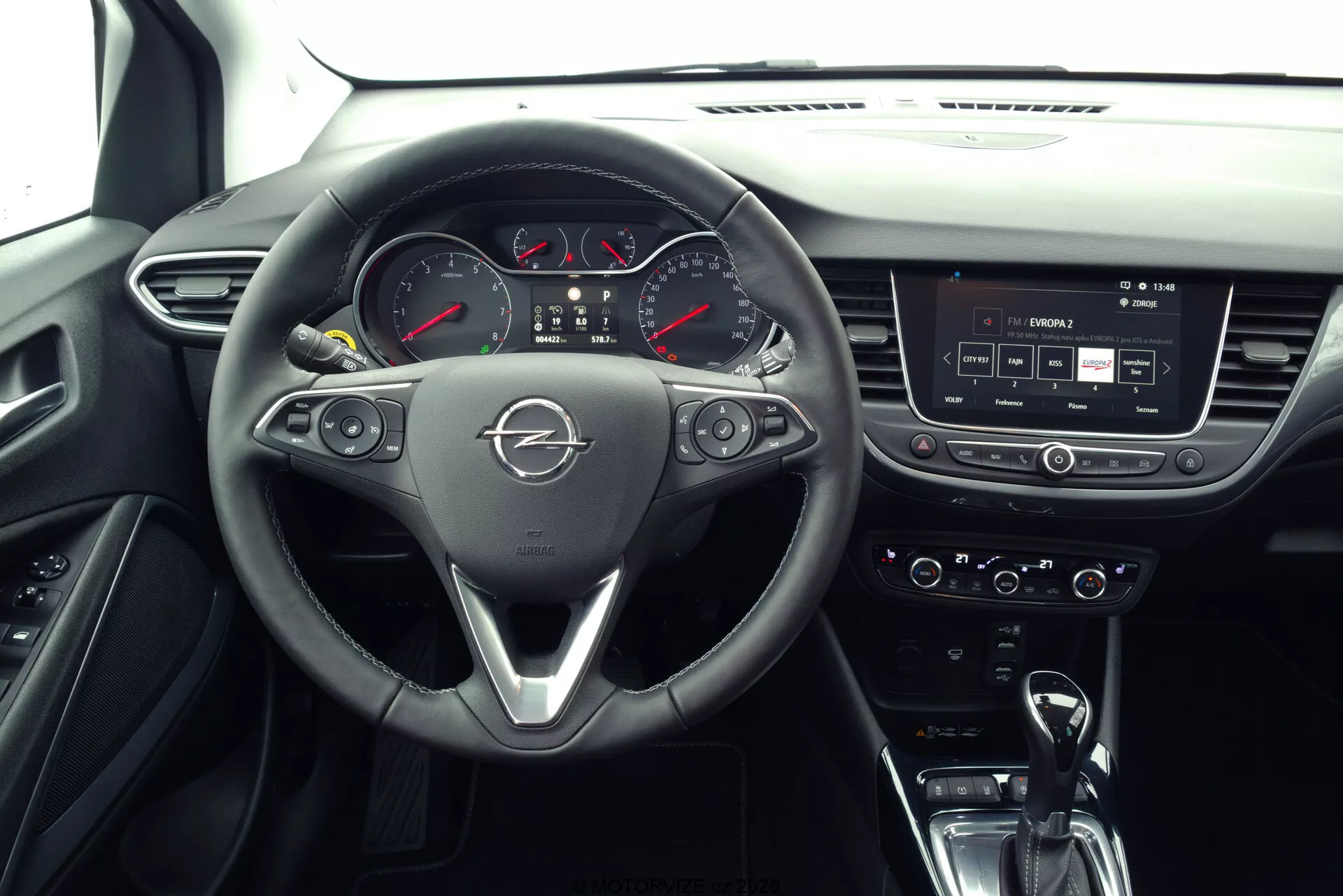 Interior of the 2021 Opel Crossland from front passenger seat view, showcasing the three-spoke leather-wrapped steering wheel with multifunctional buttons, prominently displayed brand logo, analog instrument cluster with digital display, central console with infotainment screen, climate control buttons, modern automatic transmission shifter, and contemporary interior design featuring a mix of materials including leather, plastic, chrome, and metallic accents.