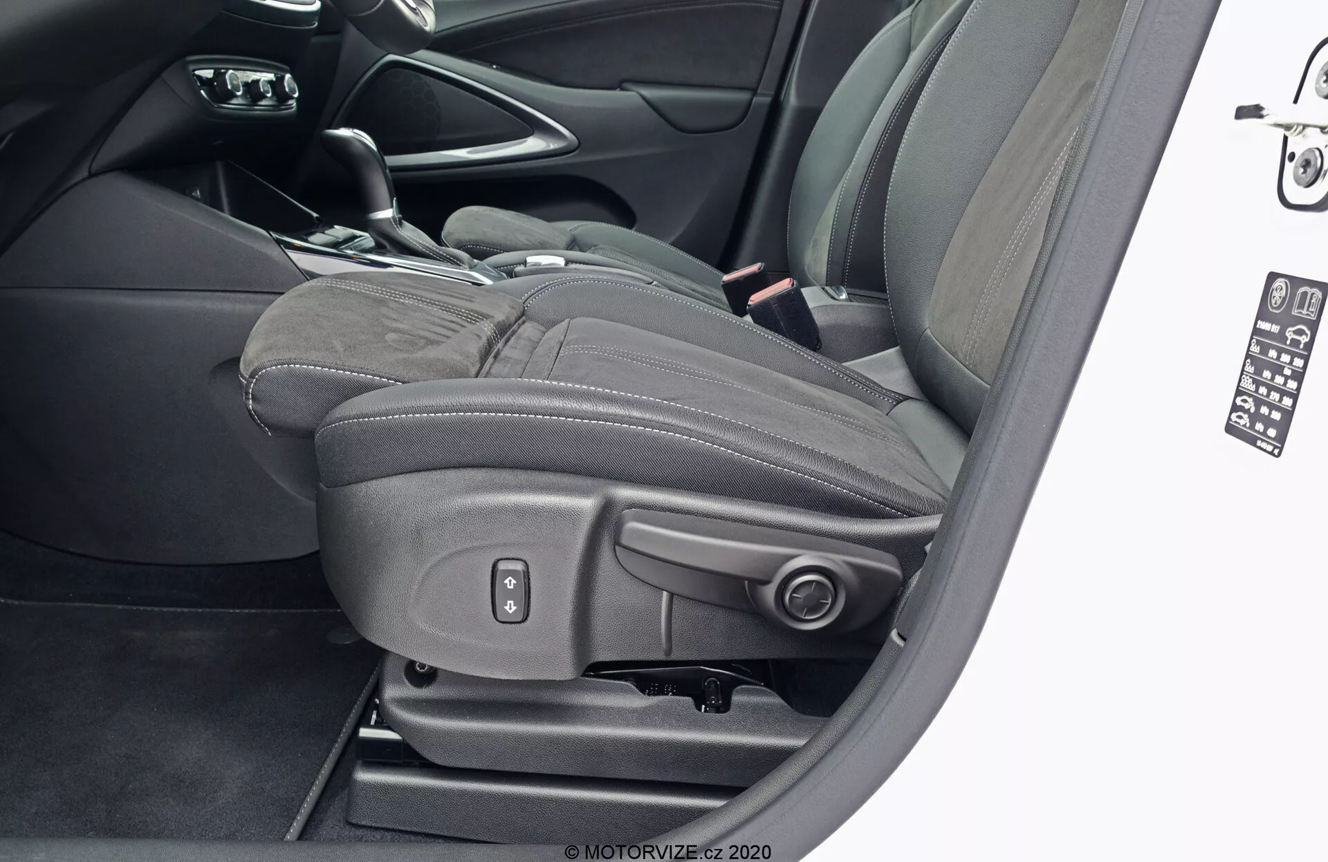 Close-up view of the driver's seat of a 2021 Opel Crossland from the exterior with the door open, showcasing black leather upholstery with prominent stitching and integrated adjustable headrest, metallic power adjustment controls for seat position, height, tilt, and lumbar support, indicating advanced comfort and luxury features.