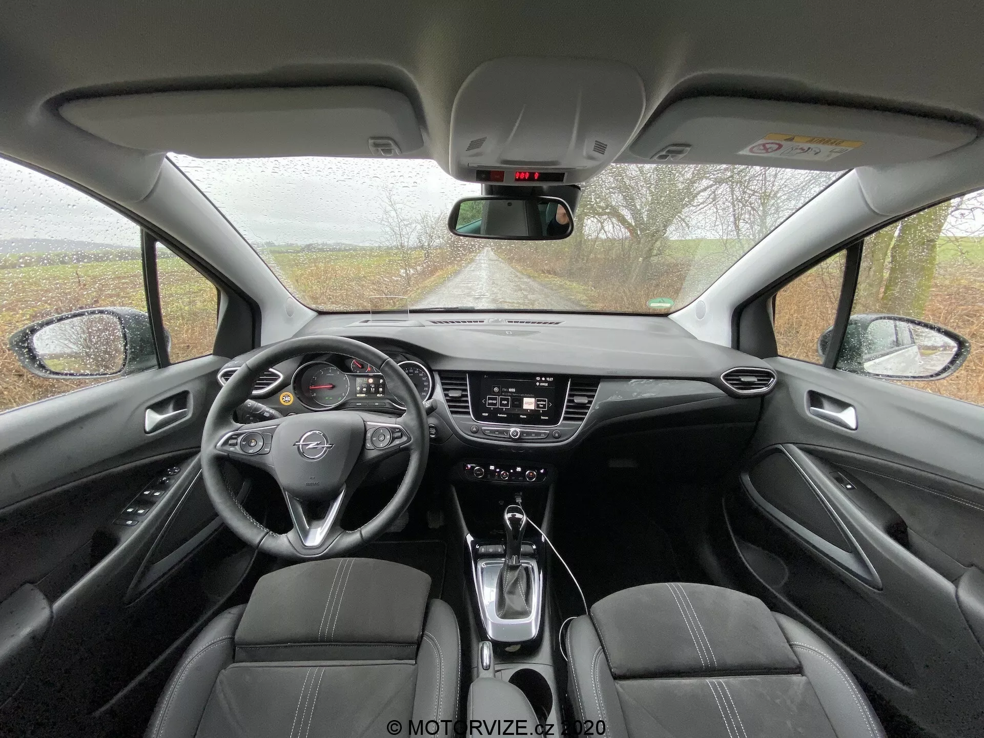 Interior view of the 2021 Opel Crossland from the rear seating area, showcasing front dashboard, three-spoke steering wheel with emblem, digital and analog instrument cluster, touchscreen infotainment system, climate control knobs and buttons, automatic transmission gear shift lever, cup holders, upholstered front seats with perforated sections, center console with armrest, manual dimming rearview mirror, A-pillars, door panels with armrests, power window controls, and possible audio system speaker grilles.
