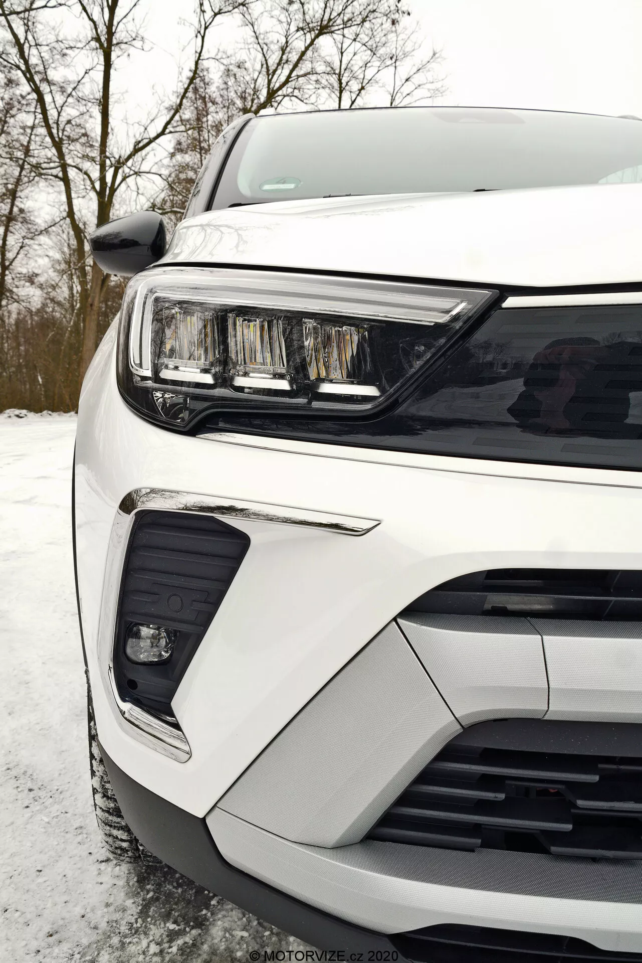 Close-up view of the front left portion of a white Opel Crossland 2021 taken from a ground-level perspective, showing the headlight assembly with a sleek horizontal LED daytime running light, a portion of the grille with horizontal slats, and the front bumper with an integrated fog light or additional driving light in a black surround and a lower cutout possibly for air intake or sensors for driver assistance systems.