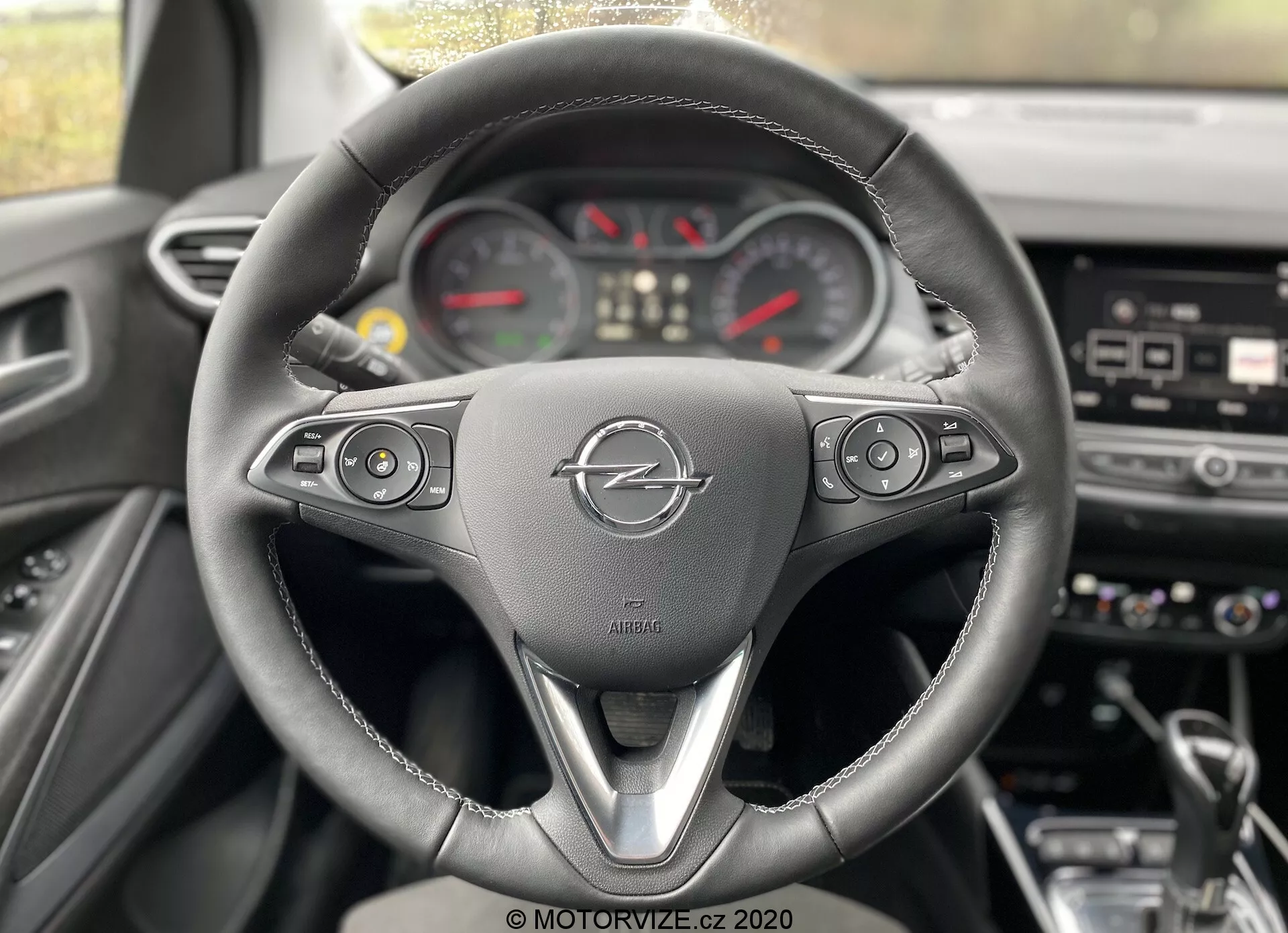 Interior of a 2021 Opel Crossland from the driver's perspective, highlighting the three-spoke steering wheel with multifunction buttons for media, cruise control, voice command, and telephone. The Opel logo and a safety airbag label are prominently displayed. Behind the wheel, a partially visible analog gauge layout includes a central speedometer, tachometer, and additional meters. The dashboard shows blurred buttons and a possible central touchscreen for the infotainment system.