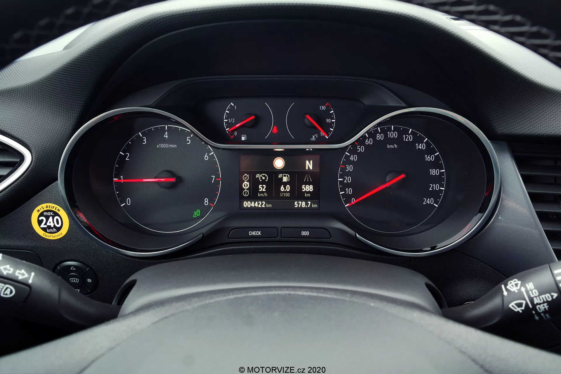 Close-up view of the instrument cluster and parts of the steering wheel in a 2021 Opel Crossland. The cluster features two primary analog dials for the tachometer and speedometer, accented with red pointers, and a central digital display showing gear position, digital speed readout, odometer, trip meter, and warning icons. The image also showcases control stalks on either side of the steering wheel, indicating technological integration in the vehicle's dashboard.