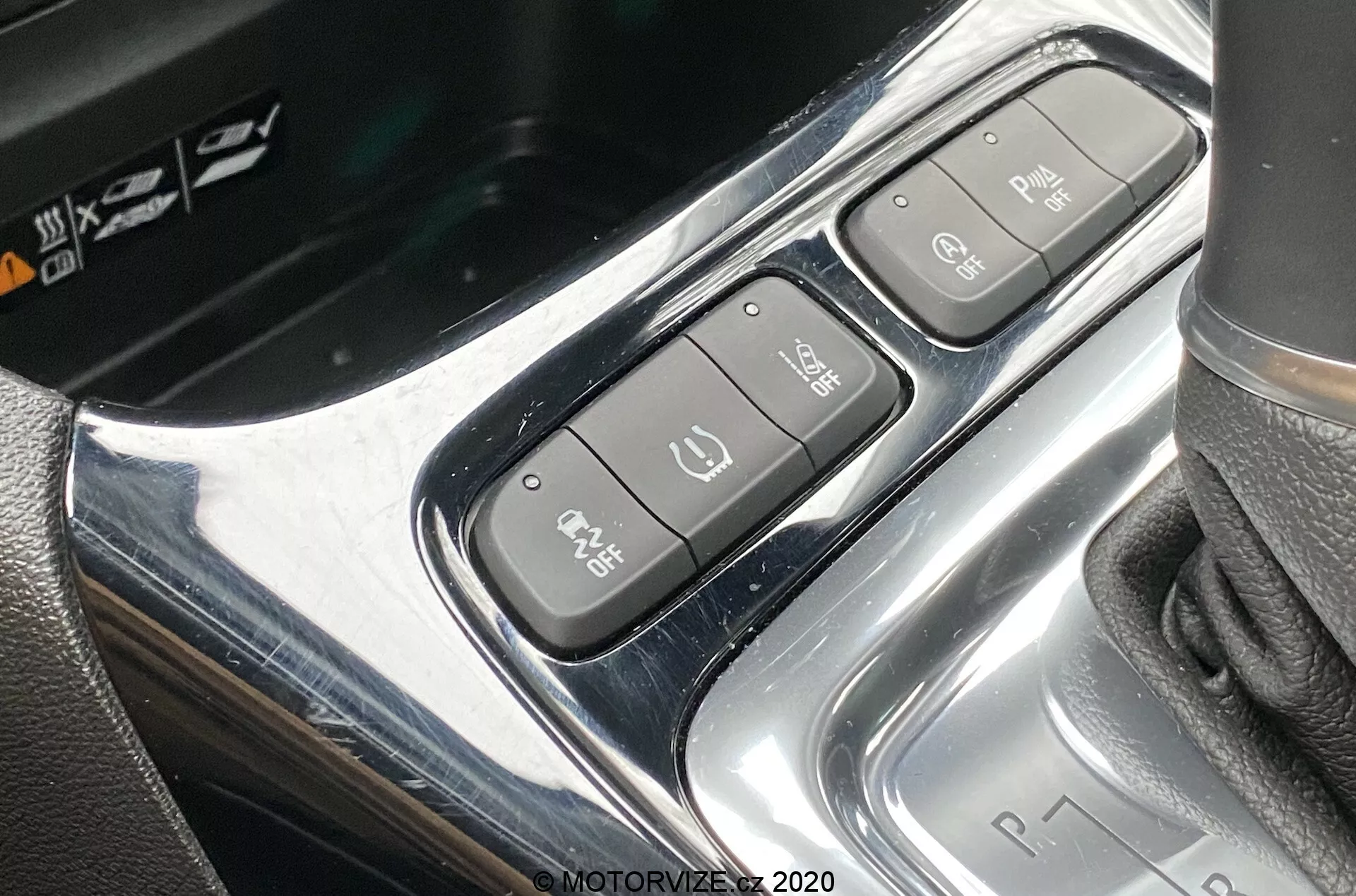 Close-up of the center console of a 2021 Opel Crossland, highlighting the control cluster near the gear shift area. The overhead, slightly angled shot shows a chrome-finished design with three distinct buttons: an electronic parking brake, Auto Hold, and Park Assist. The gear shift to the right displays a 'P R N D' sequence, indicating an automatic transmission.