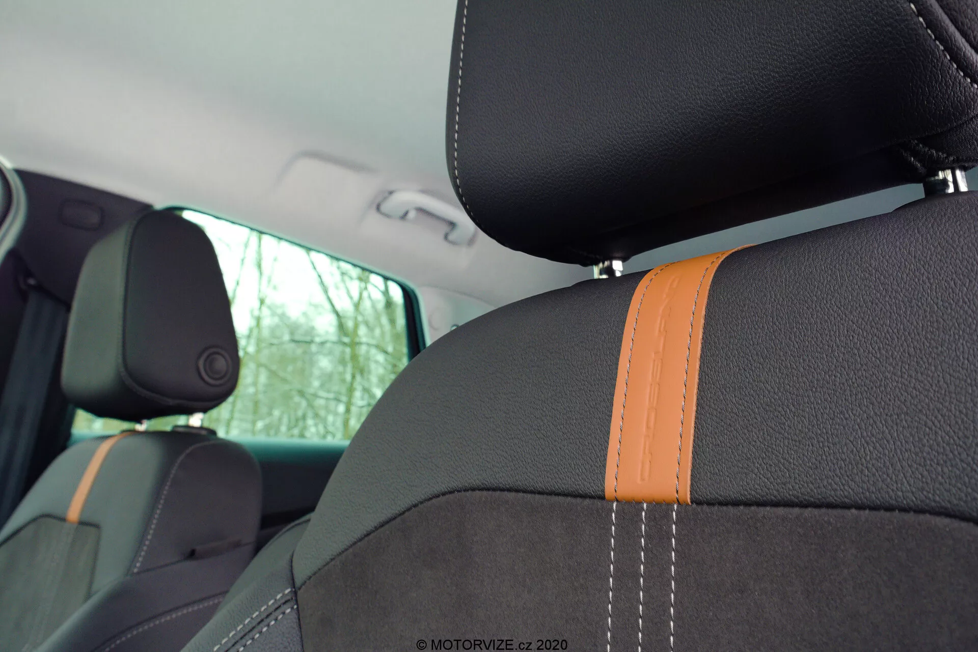 Detailed view of the Opel Crossland 2021's interior, focusing on the front and rear seats. The image is captured from the back seat perspective slightly forward and to the left, showcasing the backrest of the front passenger seat and part of the driver's seat. The seats feature dark upholstery with prominent orange stitching, suggesting a sporty theme, and integrated headrests indicative of modern design. The side bolster on the front passenger seat highlights sportiness and additional lateral support. The natural light coming through the side windows and the visible B-pillar enhance the interior's aesthetic while the watermark '© MOTORVIZE.cz 2020' in the top right corner suggests the image's origin.