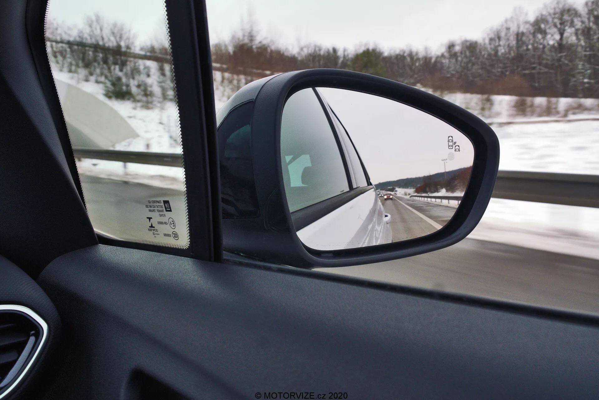 Side-view mirror of the 2021 Opel Crossland focusing on the driver's side, taken from the car's interior near the A-pillar. The mirror displays the lane beside and part of the road behind, with another vehicle visible in the snowy distance. The foreground features out-of-focus door panel elements such as window controls, highlighting interior design and safety features.