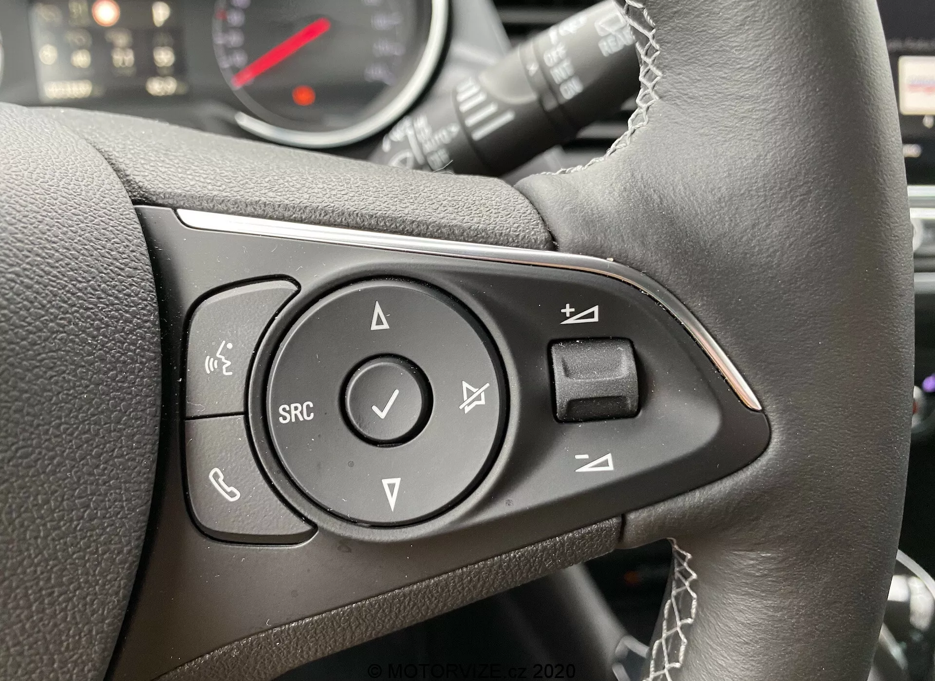 Close-up view of the multi-function steering wheel controls of the Opel Crossland 2021 from the driver's perspective, slightly shifted to the right. The black leather steering wheel features visible stitching and houses controls within a glossy black panel. The leftmost button is for hands-free calls, likely Bluetooth-enabled. Above it is a voice control button. A central directional pad with up, down, left, right arrows and an 'OK' selection button is present. Below the pad is the 'SRC' button for switching audio sources. The instrument cluster, including speedometer and possibly a tachometer, is partially visible and out of focus.
